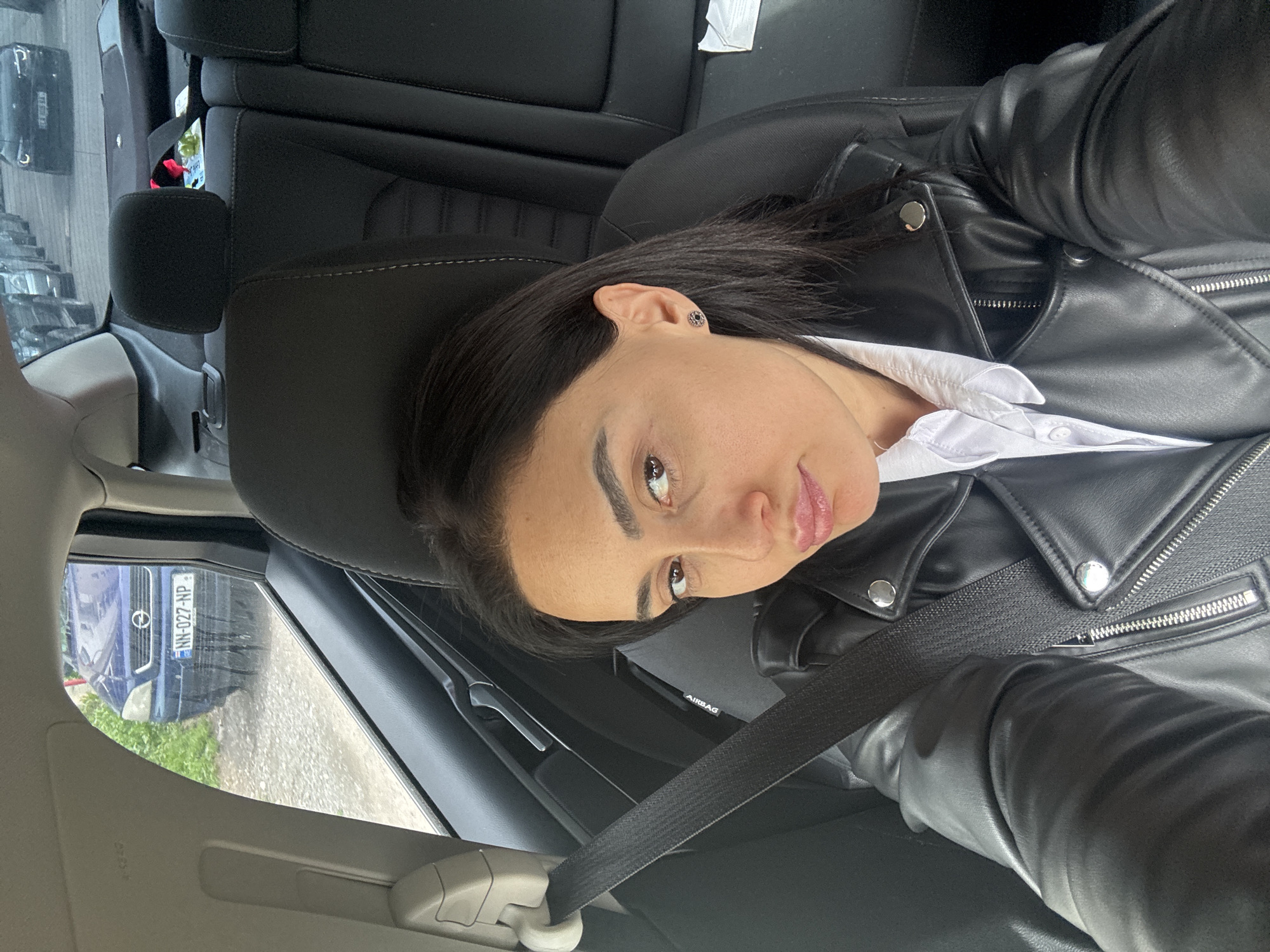 A woman is photographed from a slightly high angle inside a car. She is wearing a black leather jacket over a white collared shirt and has a seatbelt across her chest. The car’s interior is primarily black. Outside the car window, another vehicle is visible, its license plate reading "NH-027-NP". Based on the "NP" license plate designation, the location is likely Tbilisi, Georgia.