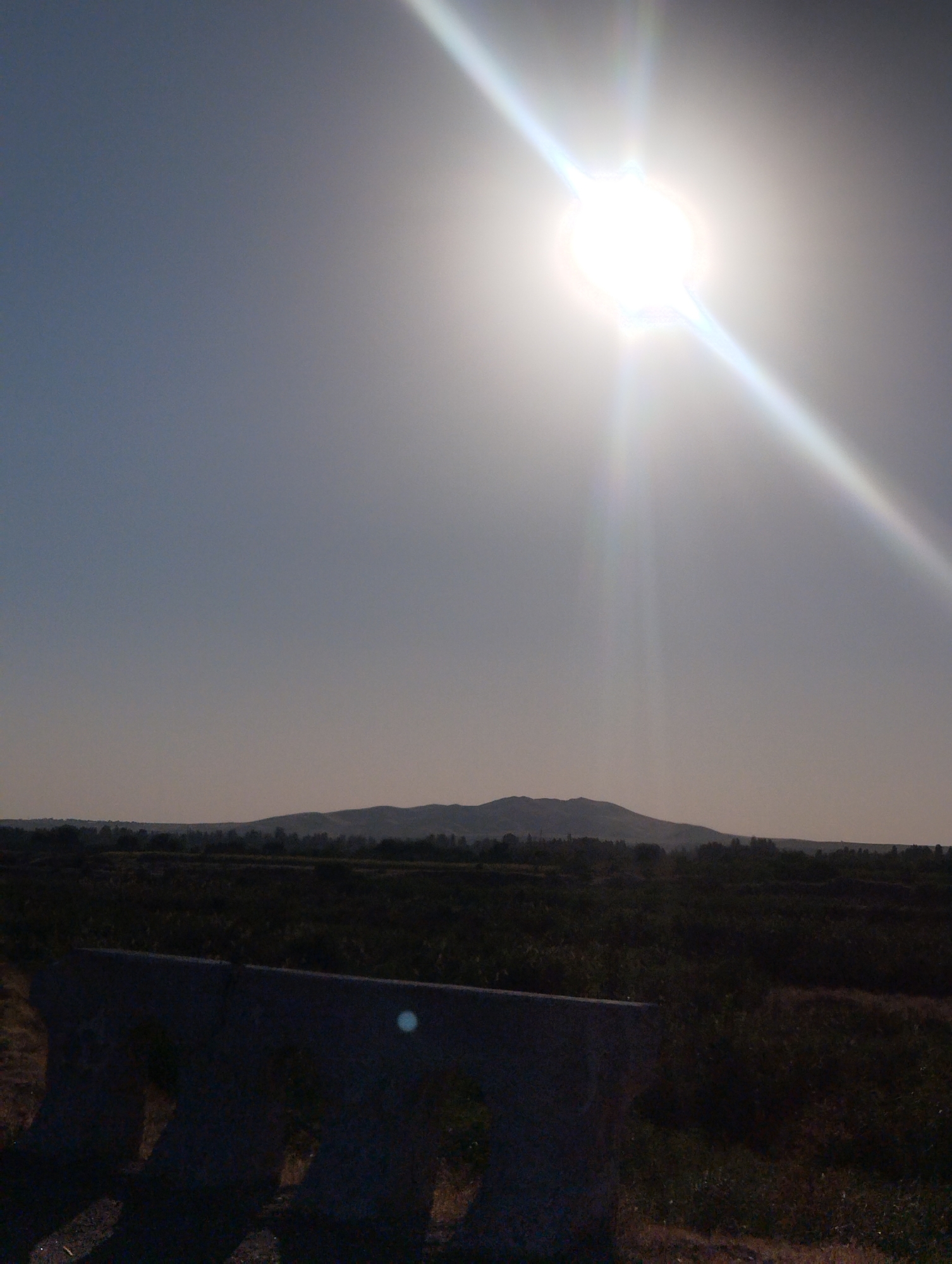 The image shows a vast expanse of sky with the sun prominently featured in the upper center, emitting bright rays of light that create lens flares. Below the sky, a distant, rolling landscape of hills is visible, silhouetted against the sky. In the foreground, a dark, textured barrier, possibly made of concrete, dominates the bottom section of the frame. The overall scene suggests an outdoor environment, likely during daylight hours, with a focus on natural elements and a somewhat stark, industrial-looking barrier.