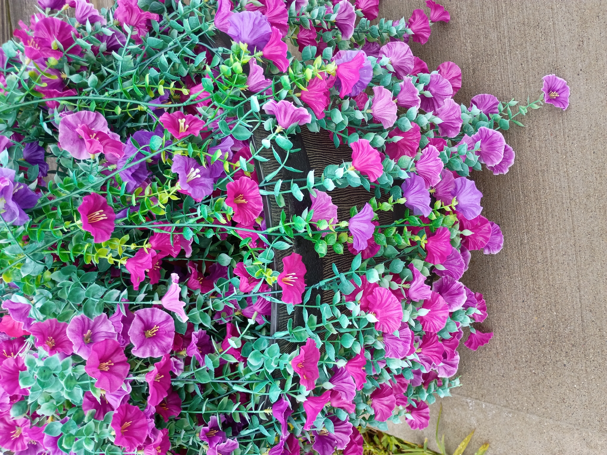 This is a close-up, overhead shot of a bundle of artificial flowers. The flowers are a mix of bright pink and deep purple, with some lighter shades of purple and pink interspersed. They are shaped like bellflowers and have delicate yellow stamens. The flowers are attached to green stems with small, oval, light green leaves. The greenery is dense, creating a lush appearance. The bundle of flowers is placed on a textured surface that appears to be concrete or stone. A dark, textured object, possibly a planter or a bench, is visible underneath a portion of the flowers. The lighting suggests it is daytime, with no harsh shadows, indicating possibly overcast conditions or indirect sunlight. The location cues suggest an outdoor setting, likely in a garden or patio area in Owingsville, United States. There is no visible text in the image. The overall impression is one of vibrant, artificial floral decoration.