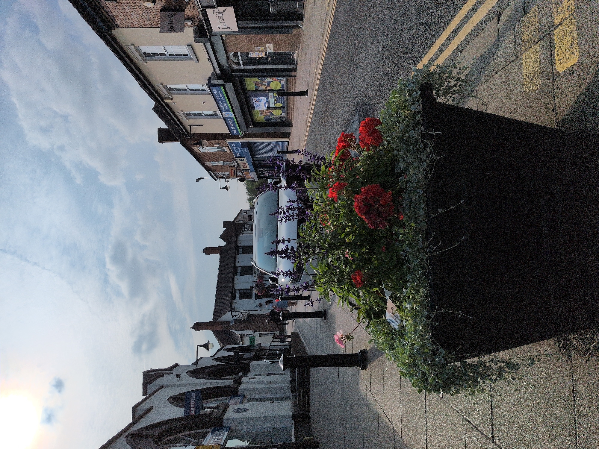 The image depicts a street scene in Dawley, United Kingdom, characterized by a row of shops on the right and a pedestrian walkway on the left. In the foreground, a large, dark planter overflows with vibrant red and purple flowers and cascading green foliage. A silver car is parked on the street, positioned behind the planter.
The buildings lining the street exhibit varied architectural styles. On the left, a white building with arched windows and a prominent "BETFRED" sign is visible. Further down the street, other shops with signs like "Clarkside" can be seen. The street itself is paved with asphalt, marked with double yellow lines. A sidewalk runs alongside the shops, separated from the street by black bollards.
Several individuals are present on the sidewalk, walking or standing, though their faces are not clearly discernible. The sky above is partly cloudy, with a bright, diffused sun suggesting it is either early morning or late afternoon. The overall impression is that of a typical high street in a British town.