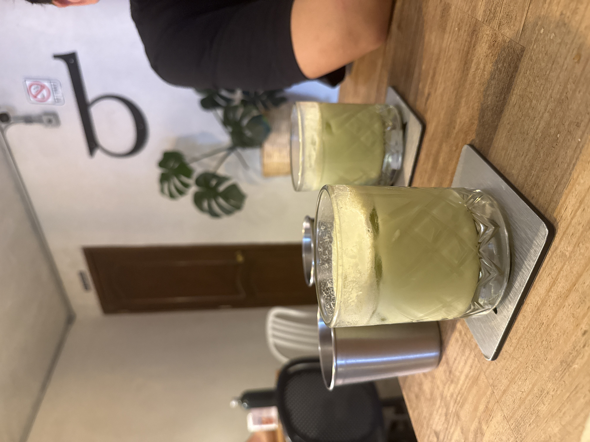 The image shows two glasses of a light green beverage, possibly matcha lattes, placed on square, silver coasters. The glasses are positioned on a light brown wooden table surface. In the background, there's a white wall with a large, stylized letter "b", accompanied by a small monstera plant. A brown door is also visible on the wall. A person wearing a black top is seated at the table. Additionally, there's a metal straw next to one of the glasses. The scene is set indoors, possibly in a cafe or restaurant. The location is San Andrés Cholula, Mexico.