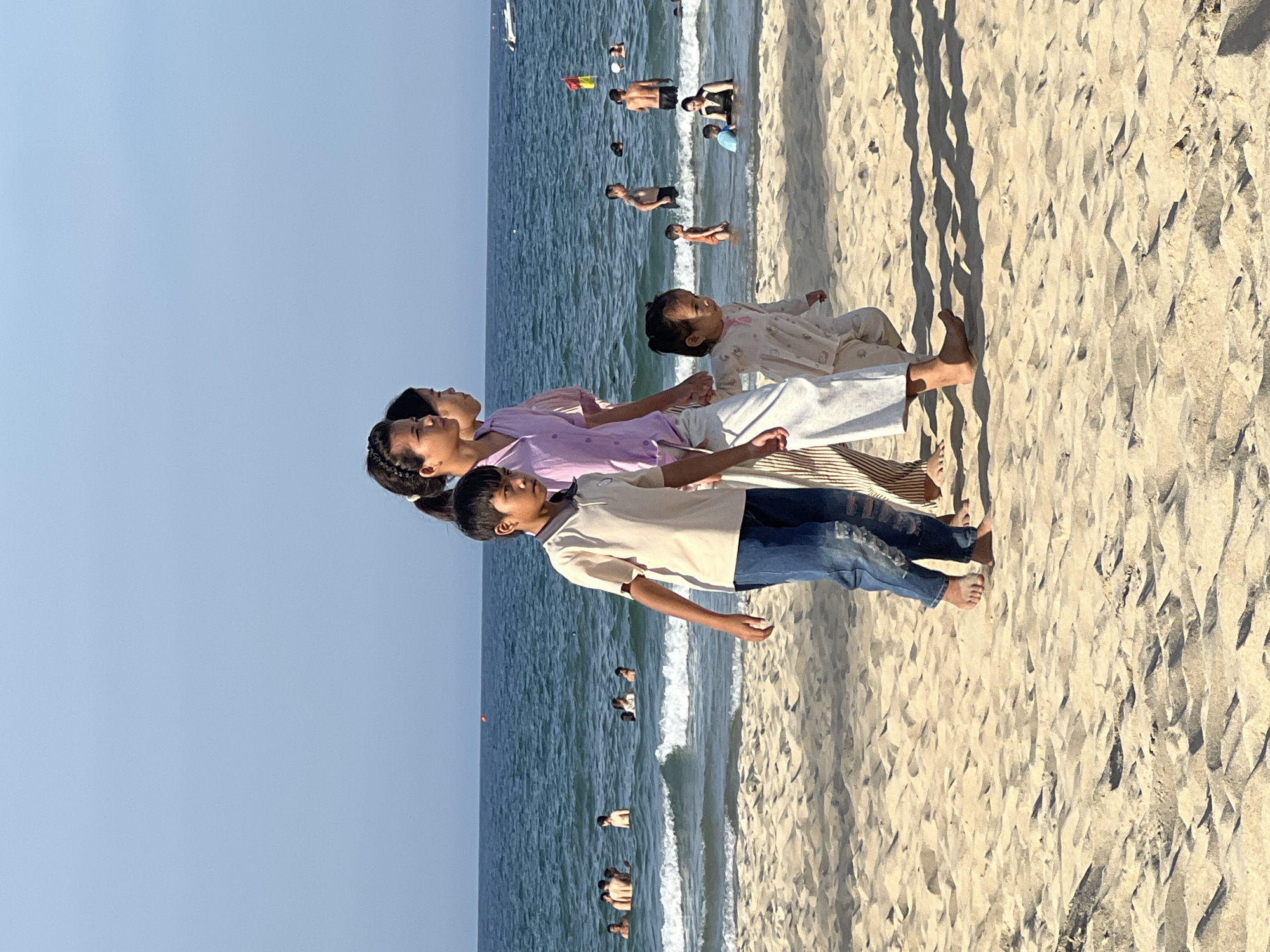 This is a full-shot, eye-level, outdoor photograph of a family walking on a sandy beach in Da Nang, Vietnam. In the foreground, four people are walking from left to right. The person furthest left is a young boy wearing a light-colored polo shirt and blue jeans. To his right is a young girl wearing a light-colored pajama set. Next to her is a young woman wearing a light purple blouse and striped pants. Behind her is another young woman, mostly obscured, wearing a light-colored outfit. They are walking on a light-tan, sandy beach with some shadows cast on it. In the background, the ocean is a deep blue with moderate waves. Several people are in the water, some swimming and others standing. A red buoy is visible in the water. The sky is a clear, light blue.