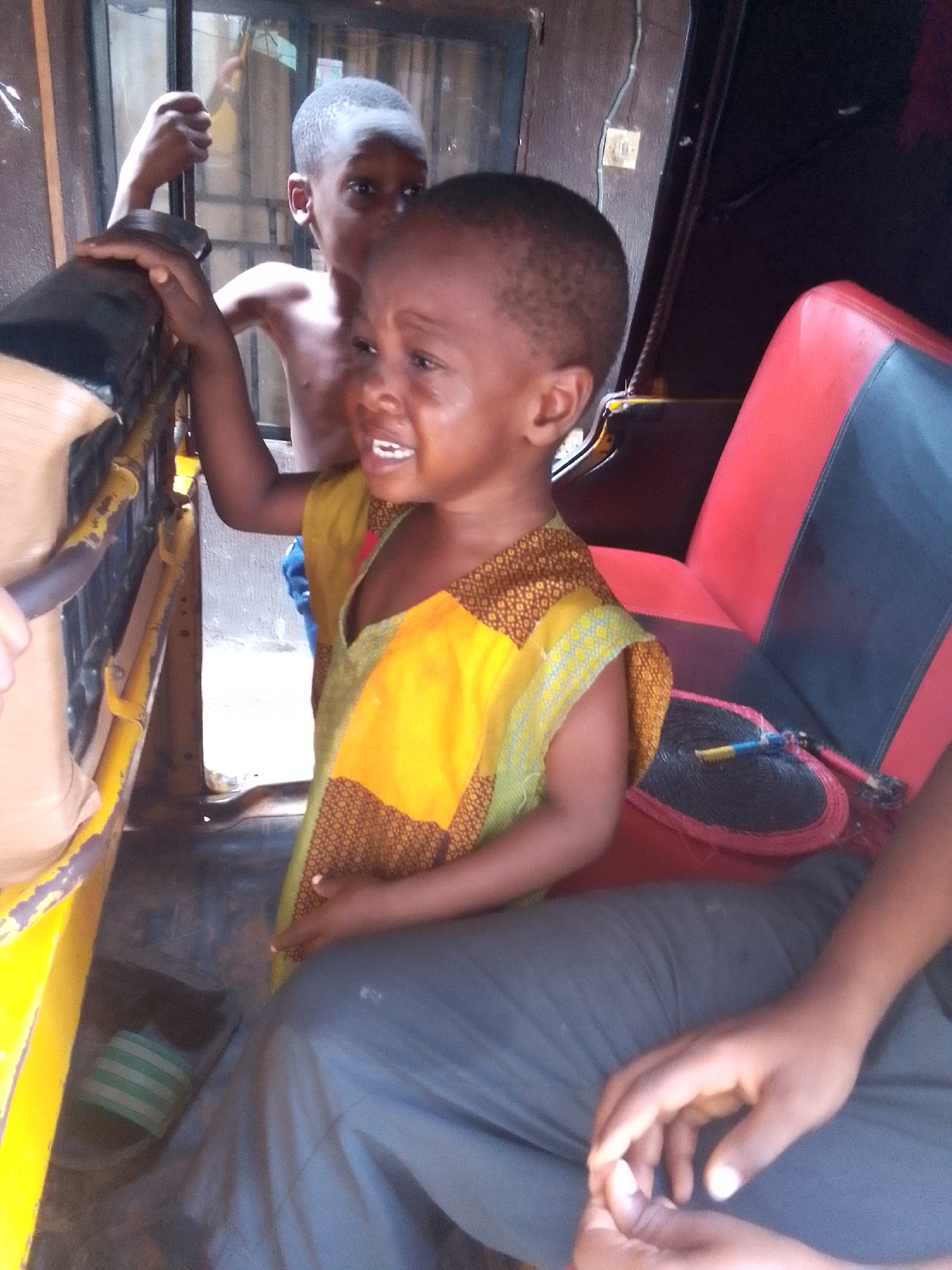 Two children and a seated person are in the interior of a vehicle in Nkpor, Nigeria. The vehicle has a red and black seat and is painted yellow. One child is shirtless and standing, holding onto the vehicle's frame, while the other child is seated, wearing a yellow and brown garment, and crying. The seated person wears grey pants. A hand fan rests on the seat next to the crying child.