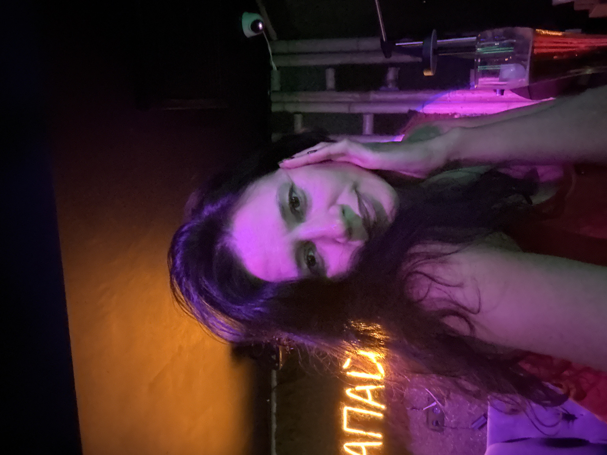 A woman with dark, wavy hair is leaning her head on her hand, looking at the camera. She is illuminated by purple and orange neon lights. In the background, a neon sign with Cyrillic lettering and a security camera are visible. A glass of beer sits on a table to the right of the woman. The environment appears to be an indoor establishment, possibly a bar or club.