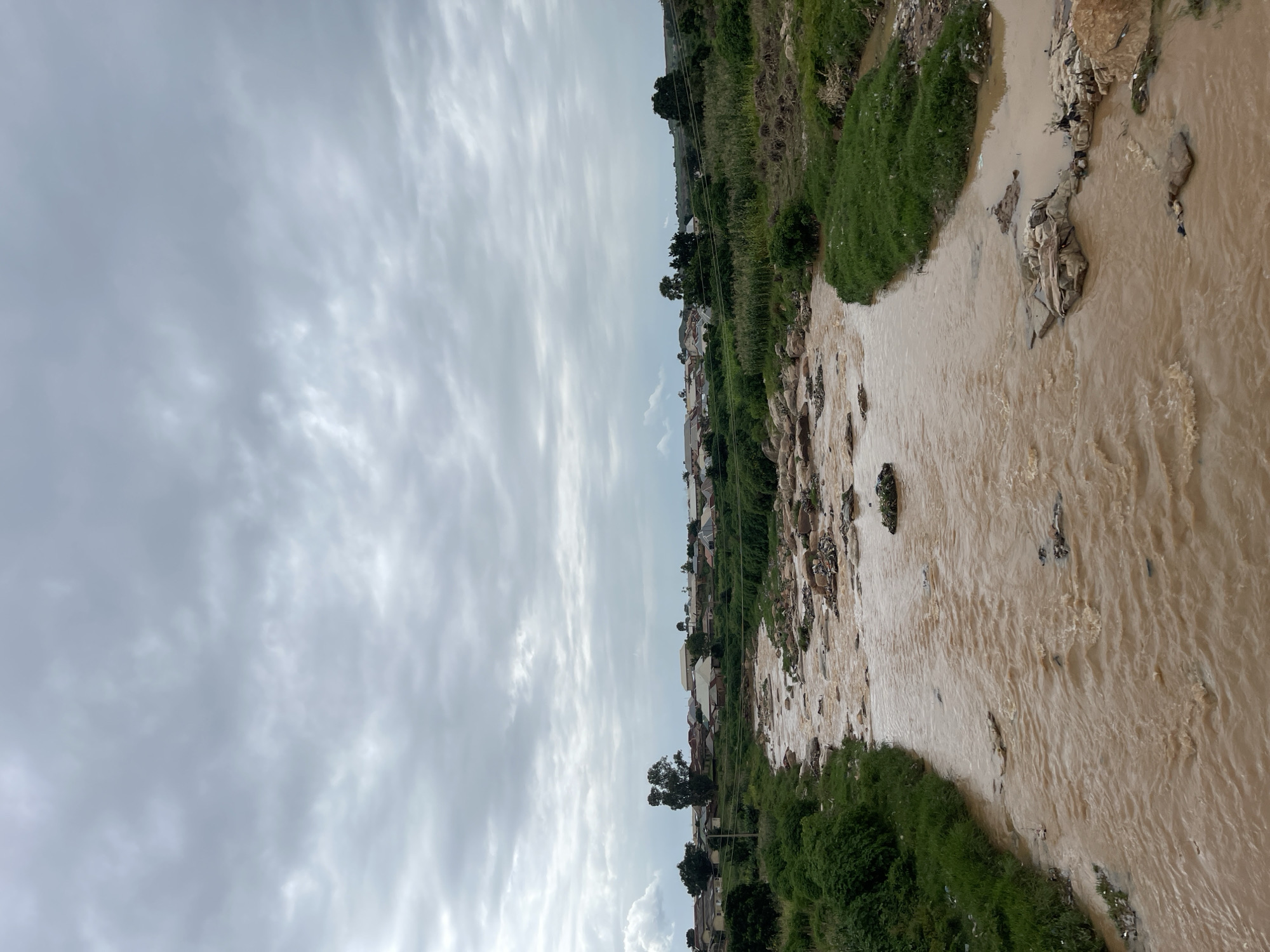 The image depicts a murky, brown river flowing through a semi-urban area. The riverbanks are lined with lush green vegetation and scattered rocks. Debris, including plastic waste and what appears to be fabric, is visible in the water and along the banks, indicating pollution. In the background, a settlement of houses with corrugated iron roofs is nestled amongst trees and greenery, suggesting a rural or peri-urban environment. The sky is overcast with heavy, grey clouds. The scene is characteristic of a polluted watercourse in a developing region, possibly in or around Rafin Par, Nigeria, given the architectural style of the houses and the natural landscape. There are no people visible in the image.