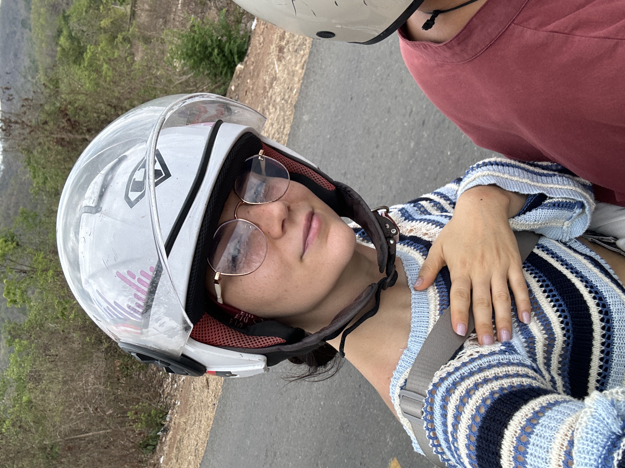 The image shows a woman wearing a white helmet with a clear visor and round glasses, sitting on what appears to be a motorcycle. She is wearing a blue and white striped open knit sweater, and a seatbelt is visible across her torso. Another person wearing a helmet and a red shirt is partially visible to the right of the woman. The background reveals a road, vegetation-covered terrain, and distant hills. The location is Hàm Thuận Bắc, Vietnam.