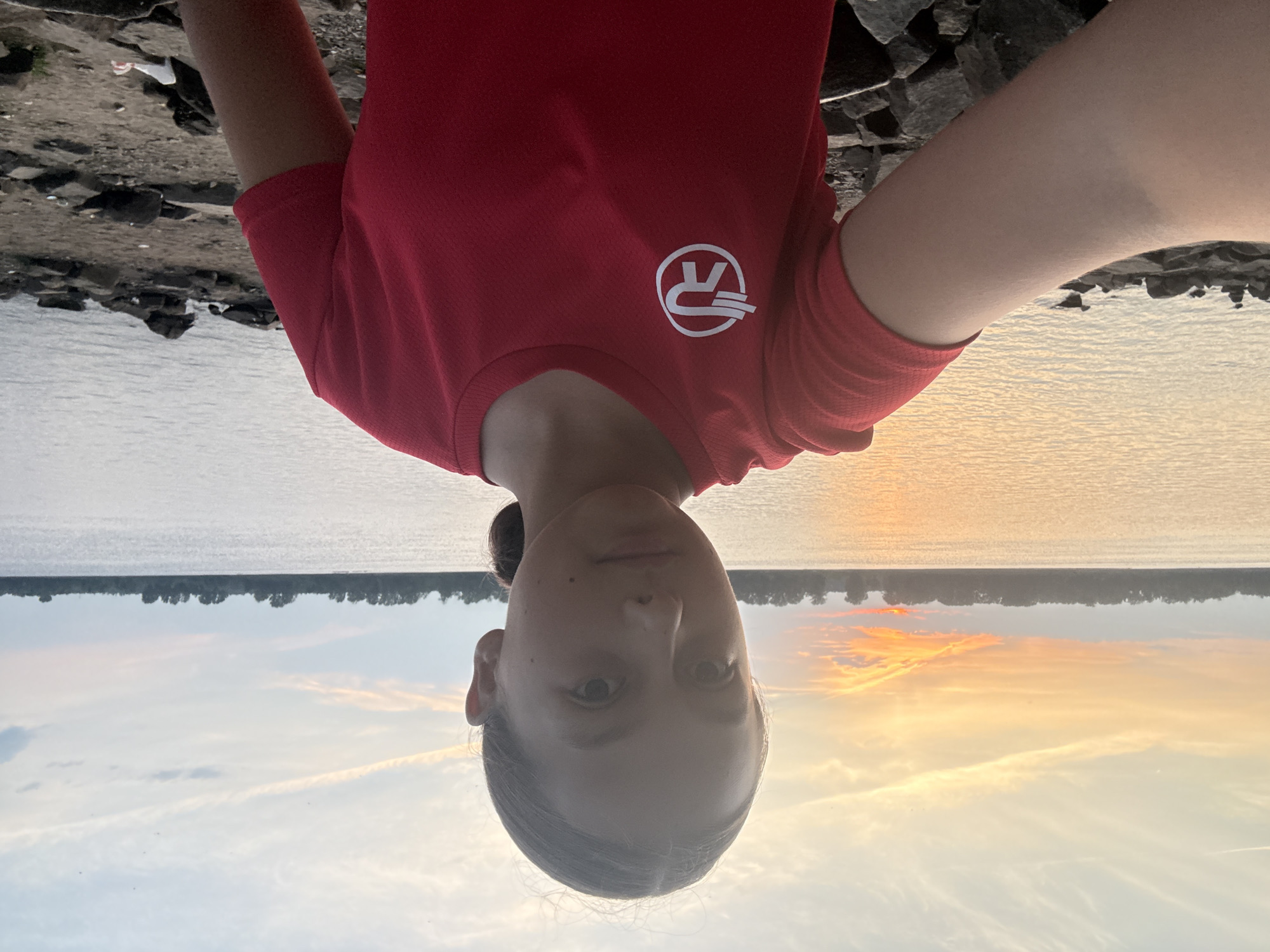 The image is a selfie taken by a young girl with her hair pulled back in a ponytail, standing outdoors by a body of water. She is wearing a red t-shirt with a white logo on the left chest. The background shows a lake or river with a tree line along the far bank and a sunset with orange and yellow hues in the sky. The ground in the foreground is rocky and sandy, with some debris visible. The location is identified as Chaikovsky, Russia.
