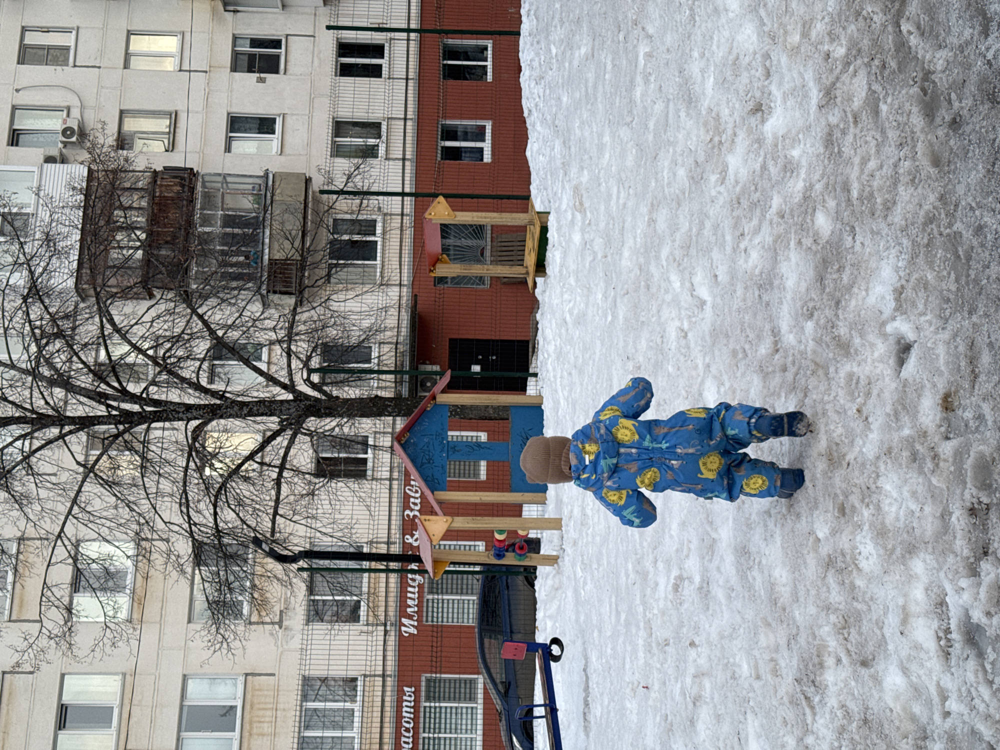 The image depicts a young child in a blue snowsuit with a brown hat walking in the snow. In the background is an apartment building with bare trees in front. A playground structure is situated in front of the building. The playground structure has text in Russian on the wall behind it. The image appears to be taken in Chaikovsky, Russia.