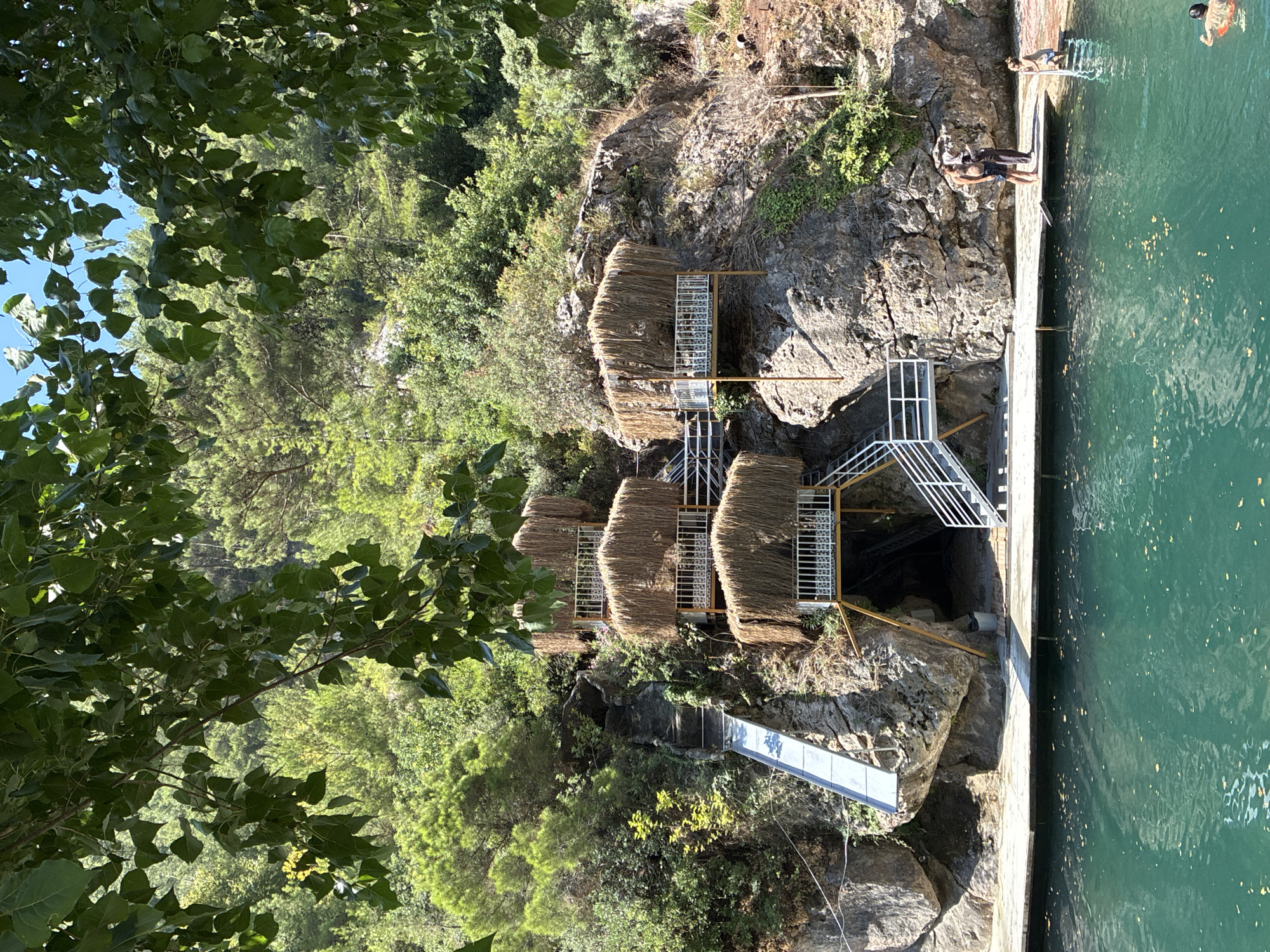 The image depicts a recreational area adjacent to a body of turquoise water, likely a natural pool or lagoon, situated within a rocky, forested environment. Multiple tiered platforms with thatched roofs are built into the rocky hillside. One platform has a slide descending to the water's edge. Stairs provide access between the platforms and to the water. On the wooden deck surrounding the water, several people are present: two individuals stand near the rocks, one appears to be speaking on a phone, while another person is holding onto a ladder extending into the water. In the water, another person is visible. The foliage suggests a Mediterranean or subtropical climate. This scene is located at the Dim River Canyon in Alanya, Turkey, a popular tourist destination known for its natural beauty and outdoor activities.