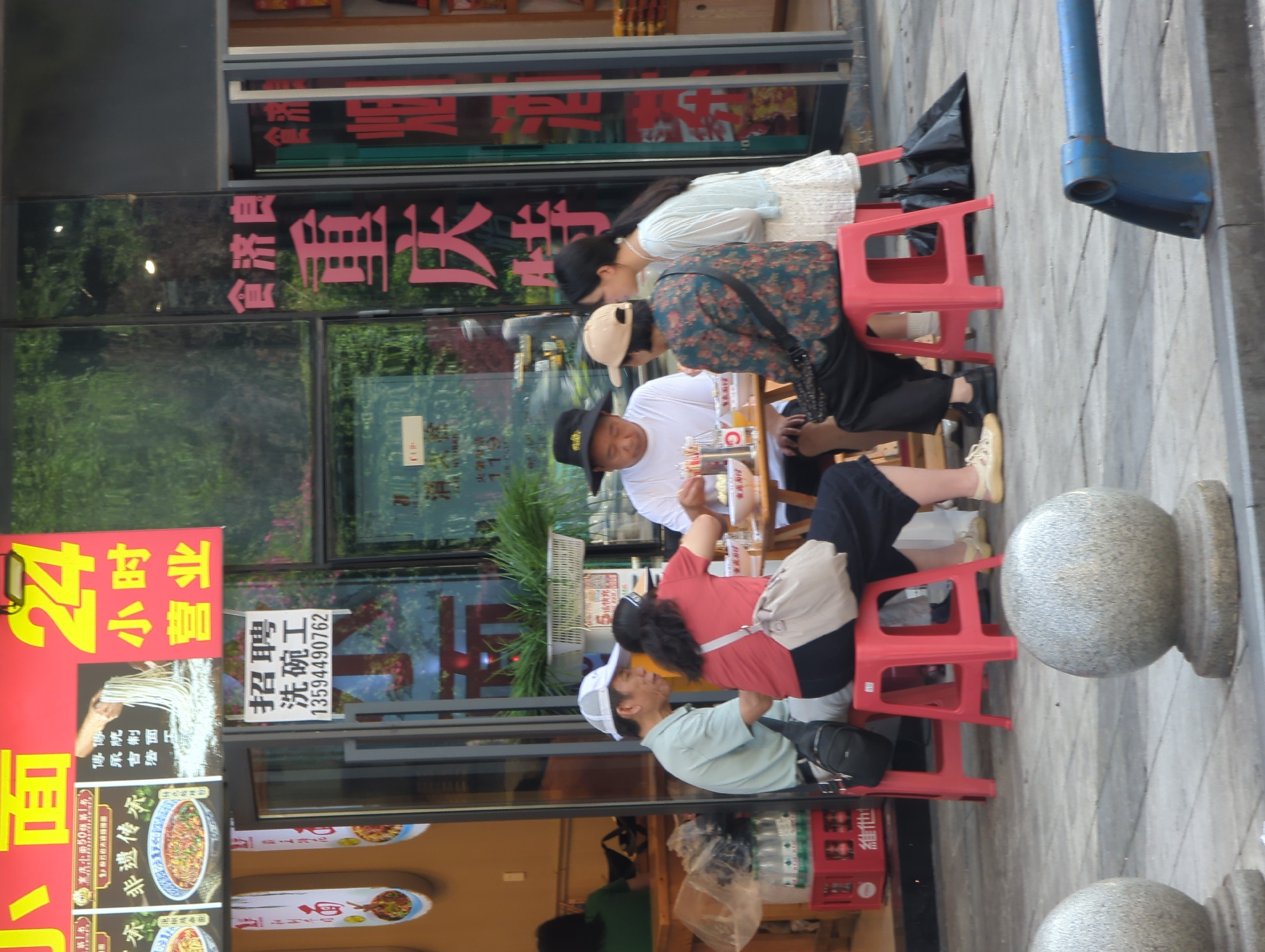 A group of four adults are gathered around a table outside a restaurant in Yuzhong District, China. Two men and two women are seated on red stools. One man is wearing a white t-shirt and a black hat, and the other is wearing a light blue polo shirt and a white baseball cap. A woman in a red shirt is sitting next to the man in the white polo. The fourth person is a woman wearing a patterned jacket and a tan baseball cap. They appear to be eating and drinking, with bowls and bottles on the table. The restaurant has a large red sign with yellow text and a glass storefront displaying menus and food items. In the background, there are more signs with Chinese characters and a glimpse of the interior of the restaurant. The sidewalk is paved with gray tiles, and there are cylindrical concrete bollards visible in the foreground.