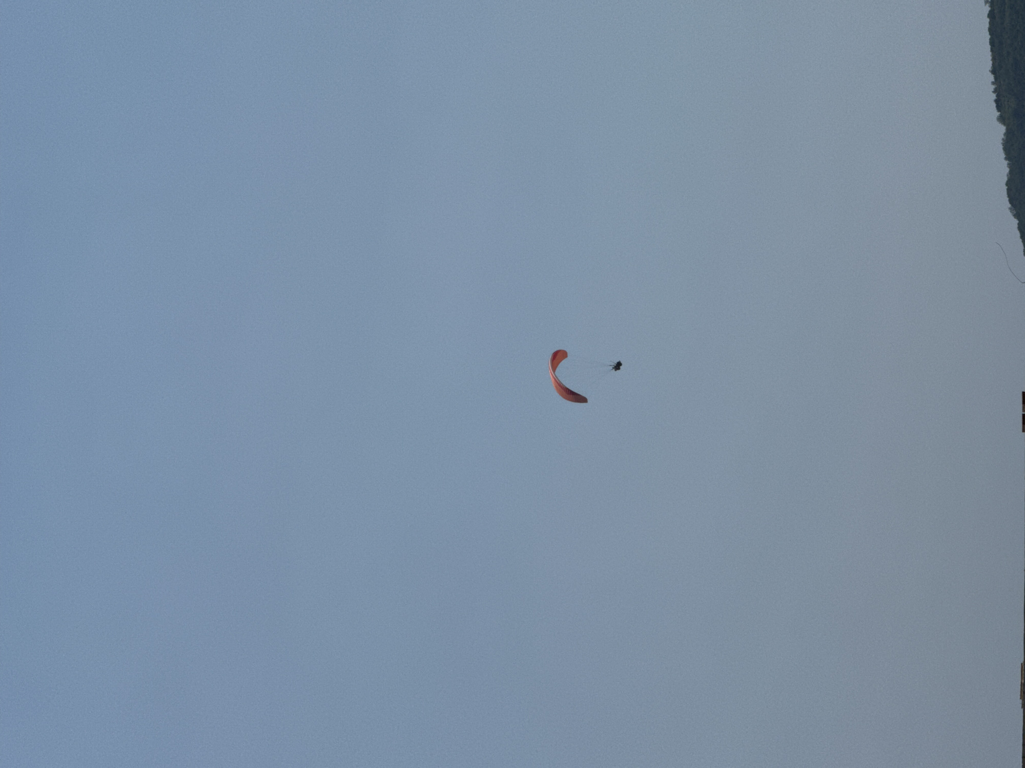 A paraglider, colored red, is suspended against a clear blue sky. The paraglider, with a person attached, is captured in mid-air. Below the paraglider, the top of a green, tree-covered hill is visible. At the very bottom of the image, a dark horizontal line suggests a distant structure or landscape element. The overall scene is an outdoor activity taking place in a natural environment, possibly near the city of Boreti, Montenegro, indicated by the presence of a hilly landscape.