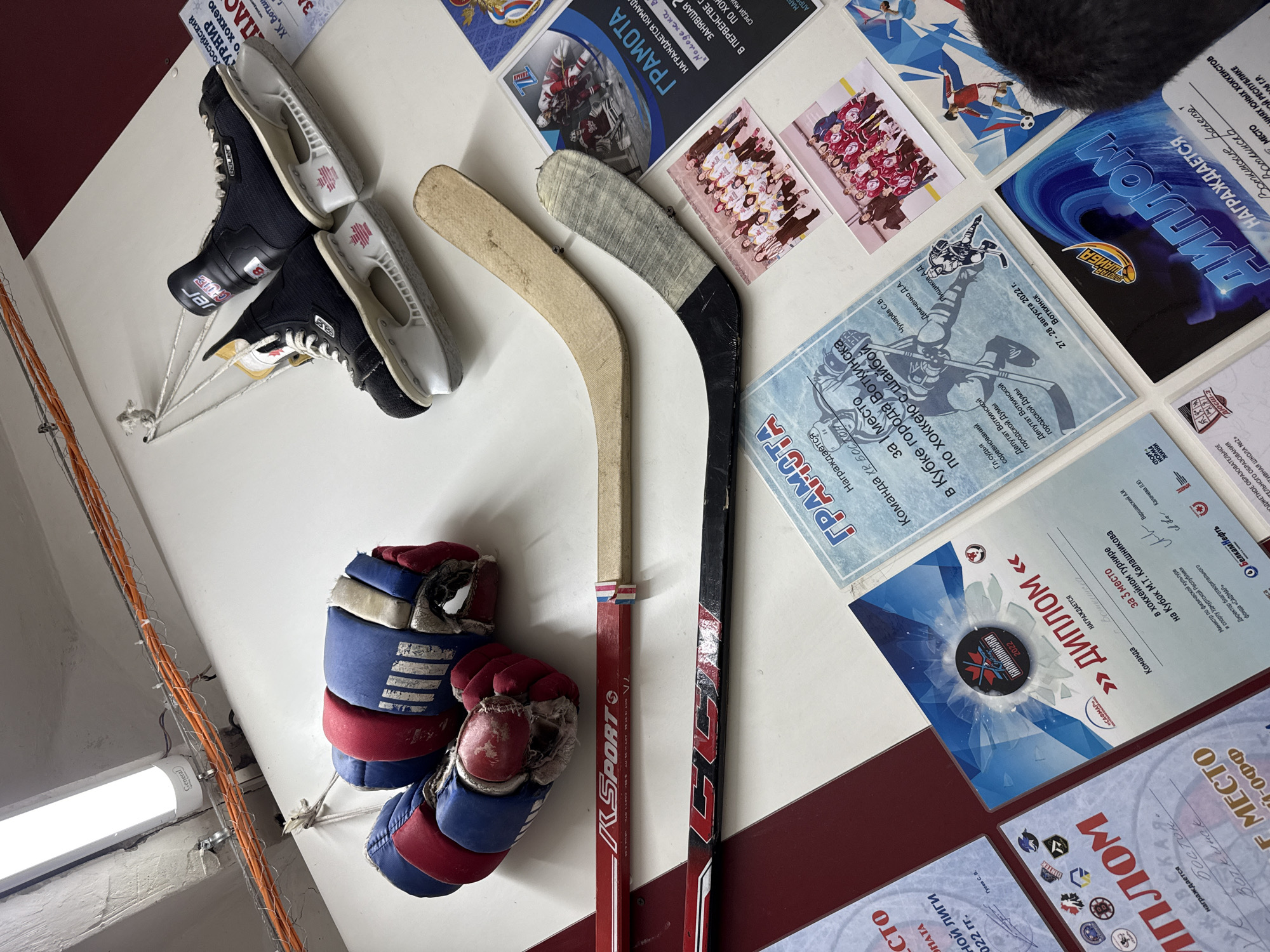 The image depicts a collection of hockey equipment and awards displayed on a white surface, likely a wall or shelf, in what appears to be a locker room or sports facility in Votkinsk, Russia. Dominating the frame are a pair of black Bauer hockey skates, suspended by white ropes. Below the skates, two hockey sticks are laid out horizontally. The upper stick is red and white with "K SPORT" branding, while the lower stick is black and white with "CCM" branding. A pair of well-worn red and blue hockey gloves are also hung, showing signs of significant use.
Scattered around the equipment are various certificates and photographs. Several "GRAMOTA" (diploma/certificate) documents are visible, some bearing the text "НАГРАЖДАЕТСЯ" (awarded to) and mentioning "Команда" (team) and "за 3 место" (for 3rd place). One of these certificates is specifically dated "27 - 28 августа 2022 г." and mentions "Кубок города Воткинска по хоккею с шайбой" (Votkinsk City Cup for Ice Hockey). Another certificate, a "ДИПЛОМ" (diploma), is also present, stating "НАГРАЖДАЕТСЯ" and also referring to Votkinsk. Several photographs show groups of young people in hockey uniforms, presumably the teams that received these awards.
The environment appears to be functional, with electrical conduits visible along the ceiling and a fluorescent light fixture overhead. The overall impression is one of accomplishment and recognition within the context of youth ice hockey in Votkinsk.