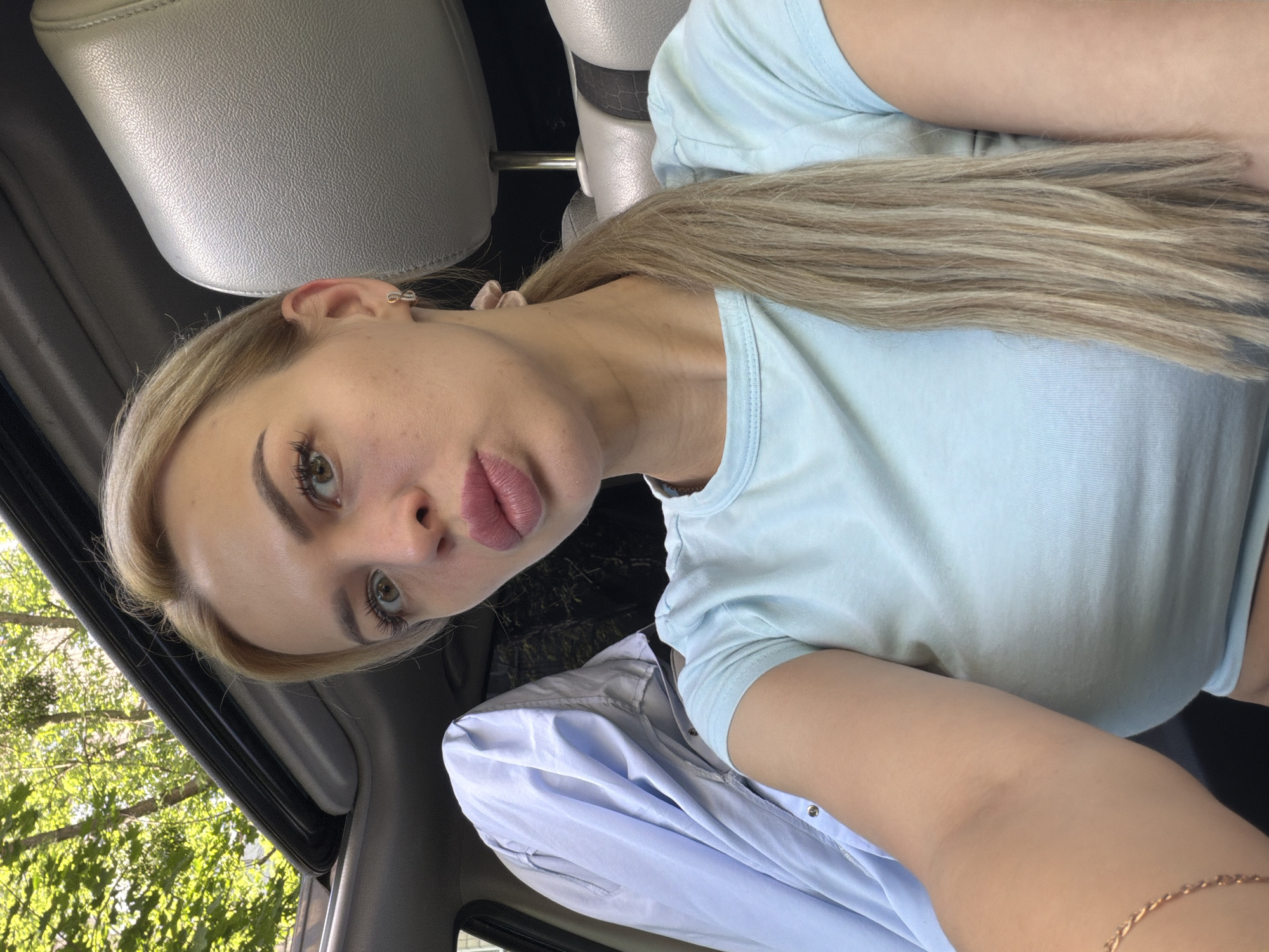 A young woman with blonde hair tied in a ponytail, wearing a light blue t-shirt, is taking a selfie inside a car. The car's sunroof is open, revealing green trees and foliage overhead. The woman is looking at the camera with her lips pursed. A light blue shirt is draped over the back seat on the left side of the image. The interior of the car appears to be gray leather. The scene is outdoors, likely during the daytime, with natural light illuminating the woman and the car's interior. There is no identifiable indication of the specific location within Kyiv, Ukraine, but the presence of trees and foliage suggests an area with greenery, possibly a park or residential street.