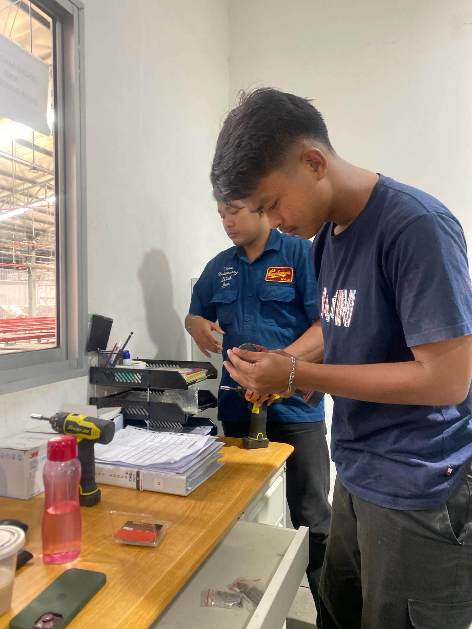Two young men are working at a wooden desk inside a brightly lit industrial workshop in Batam, Indonesia. The man in the foreground, wearing a navy blue t-shirt with a graphic print, is holding a cordless drill and appears to be assembling or repairing something. His gaze is focused on the object in his hands. Beside him, another man in a navy blue collared shirt with patches is also looking down, seemingly observing the task or assisting. The desk is cluttered with various items, including paperwork, a pink water bottle, a yellow and black cordless drill, a cell phone, and a small container with red objects. In the background, a large window reveals a glimpse of the workshop's interior, with exposed ceiling structures and red-colored equipment. The overall environment suggests a workspace where hands-on tasks are performed.