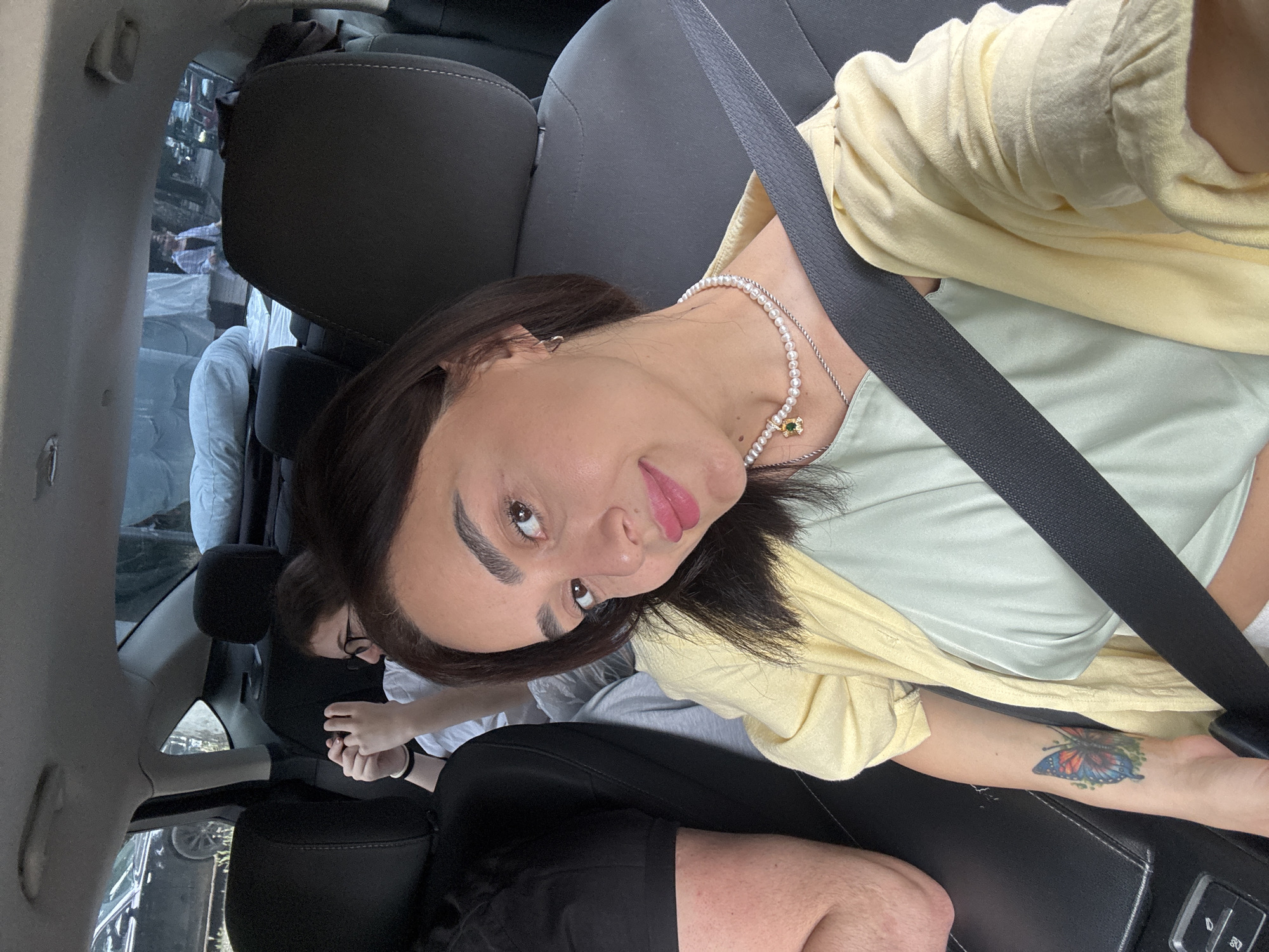 This image shows a group of people inside a car, appearing to be in motion. The primary subject is a woman in the foreground, taking a selfie. She has dark hair and is wearing a light yellow shirt over a light green top and a pearl necklace. A tattoo of a butterfly is visible on her left forearm. She is wearing a seatbelt.
In the background, other individuals are visible. To the left, a person's arm and shoulder are seen, wearing a black t-shirt. Further back, a young woman with glasses is looking at her phone. Beyond them, the rear window of the car shows a street scene with buildings and a person walking.
The car's interior is visible, including black leather seats and the seatbelt mechanism. The exterior scene visible through the windows suggests an urban environment, possibly a street in Tbilisi, Georgia, given the architectural styles of the buildings and the general ambiance. The lighting suggests it is daytime.