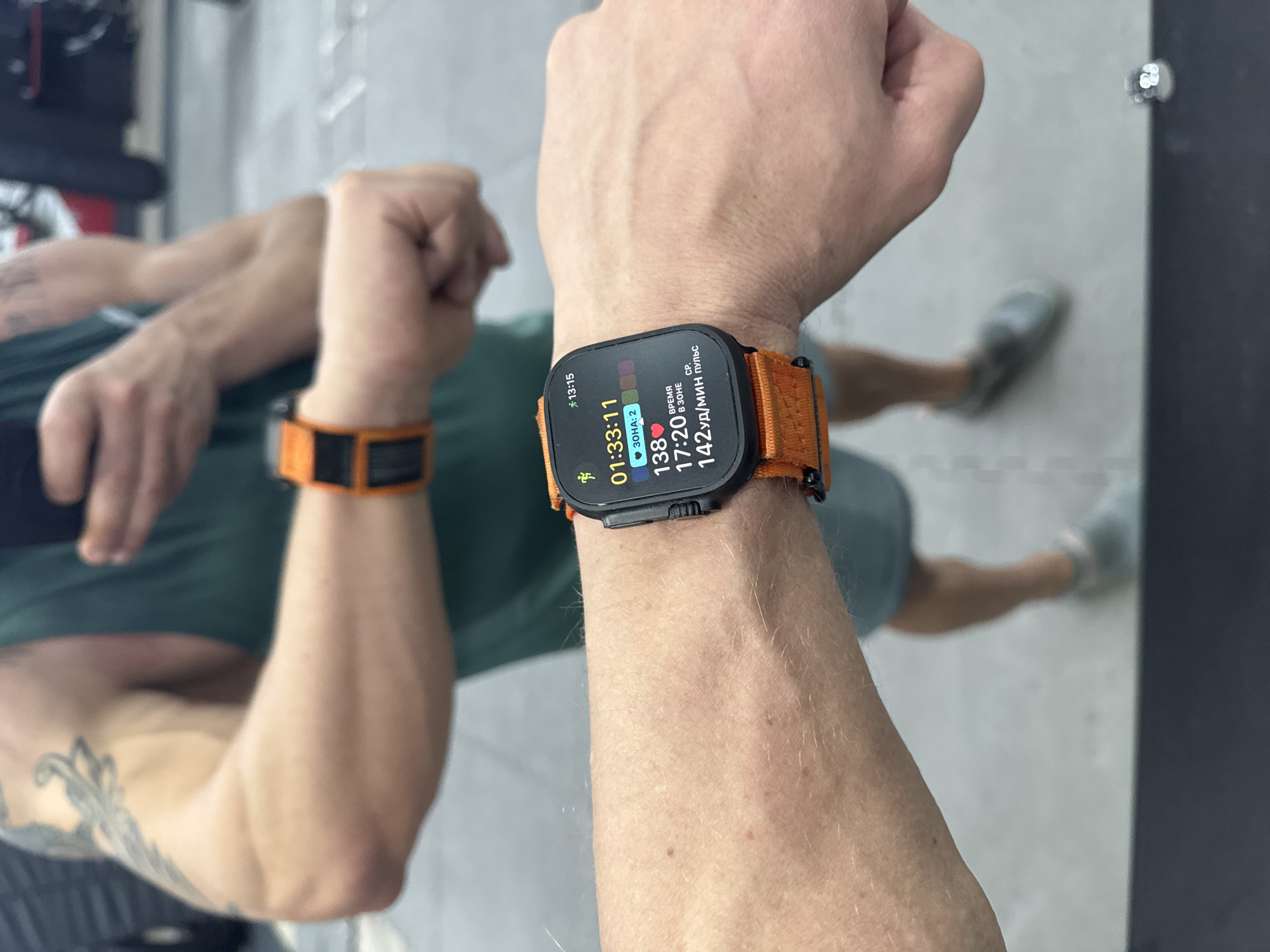 The image displays a close-up of a person's right arm, adorned with an Apple Watch Ultra featuring a striking orange fabric strap. The scene appears to be an indoor gym or fitness facility, located in Volzhsky, Russia, as evidenced by the clear reflection of the person's upper body and legs in a large mirror.
The smartwatch's screen is actively displaying workout statistics. The current time is "13:15". The workout duration shown is "01:33:11" (1 hour, 33 minutes, 11 seconds). The person's heart rate is "138" beats per minute, categorized as "3OHA: 2" (Zone: 2). They have spent "17:20 B ЗОНЕ" (17 minutes, 20 seconds in that heart rate zone), and the average heart rate for the session is "CP. Пульс 142 уд/мин" (Average Pulse 142 beats per minute).
In the reflection, the person is seen from behind, wearing a dark green or teal t-shirt. Their left arm, also visible in the reflection, sports another orange band or strap, and a tattoo is discernible on the bicep area. Light-colored sneakers and flooring, possibly concrete or gym mats, are visible towards the bottom of the reflection. The image captures a moment during an active exercise session, highlighted by the detailed workout data on the smartwatch.