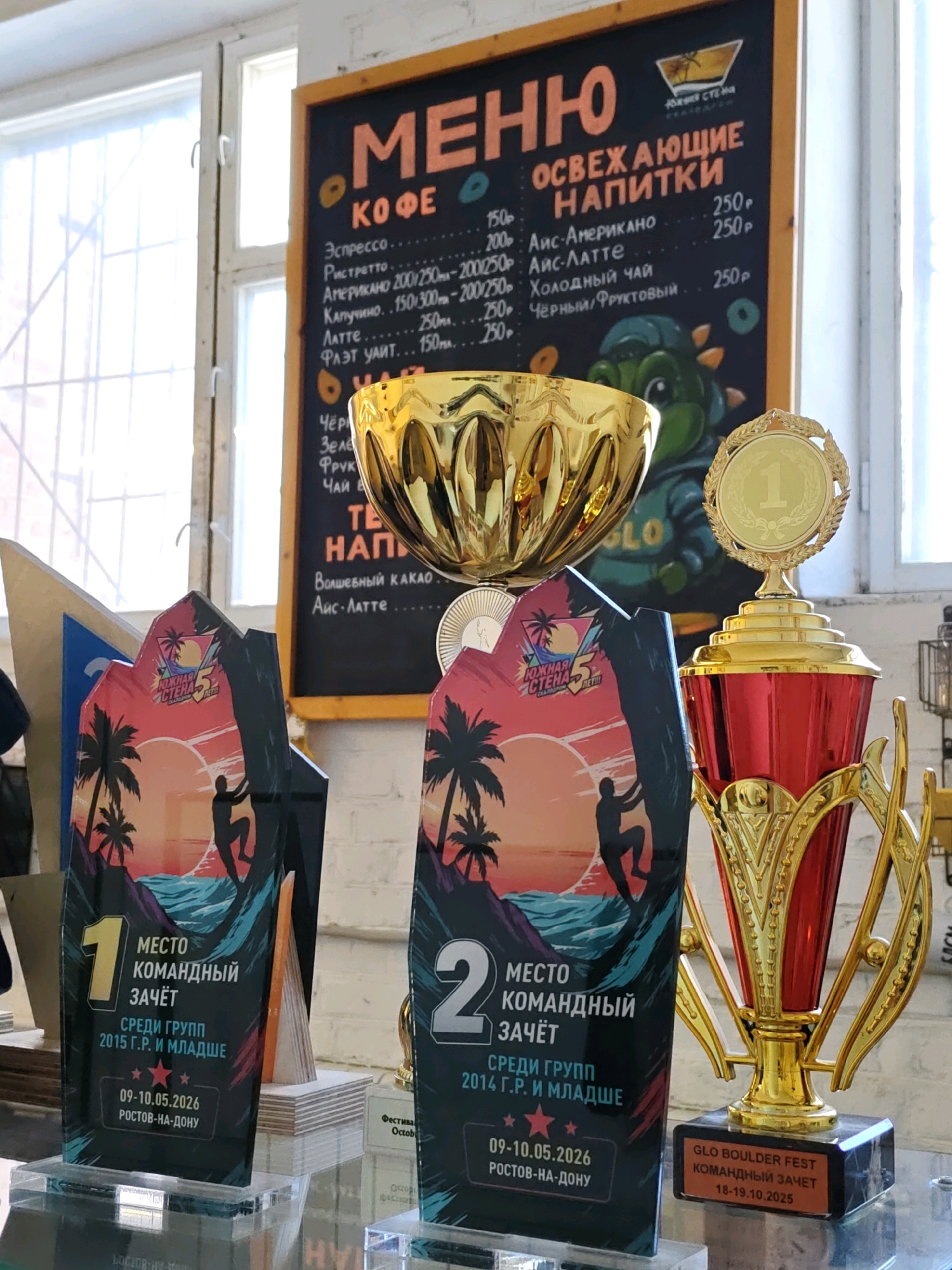 The image shows a display of awards and a menu board in what appears to be an indoor venue. The menu board, written in Russian, lists "КОФЕ" (Coffee) and "ОСВЕЖАЮЩИЕ НАПИТКИ" (Refreshing Drinks). Among the coffee options are Espresso, Americano, Cappuccino, Latte, and Flat White, with prices listed in rubles. The refreshing drinks section includes Iced Americano, Iced Latte, Cold Tea, and Black/Fruit Tea.
The awards are prominently displayed. Two of them are large, flat, and trophy-shaped with graphics depicting a rock climber silhouetted against a sunset with palm trees. These awards are labeled in Russian, with one indicating "1 МЕСТО КОМАНДНЫЙ ЗАЧЁТ" (1st Place Team Event) and the other "2 МЕСТО КОМАНДНЫЙ ЗАЧЁТ" (2nd Place Team Event). Both specify "СРЕДИ ГРУПП" (Among Groups) and then a year range, followed by "РОСТОВ-НА-ДОНУ" (Rostov-on-Don). The dates on these awards are "09-10.05.2026".
A large, traditional golden trophy with red accents stands to the right of the flat awards. Its base has a plaque that reads "GLO BOULDER FEST КОМАНДНЫЙ ЗАЧЕТ 18-19.10.2025", also indicating a location in Rostov-on-Don.
In the background, a golden bowl-shaped object rests on the menu board, and a graphic of a cartoon dinosaur is visible. The environment suggests a cafe, sports venue, or an event space, likely a climbing-related establishment given the boulder fest mentioned. The lighting suggests an indoor setting with natural light coming from windows on the left.