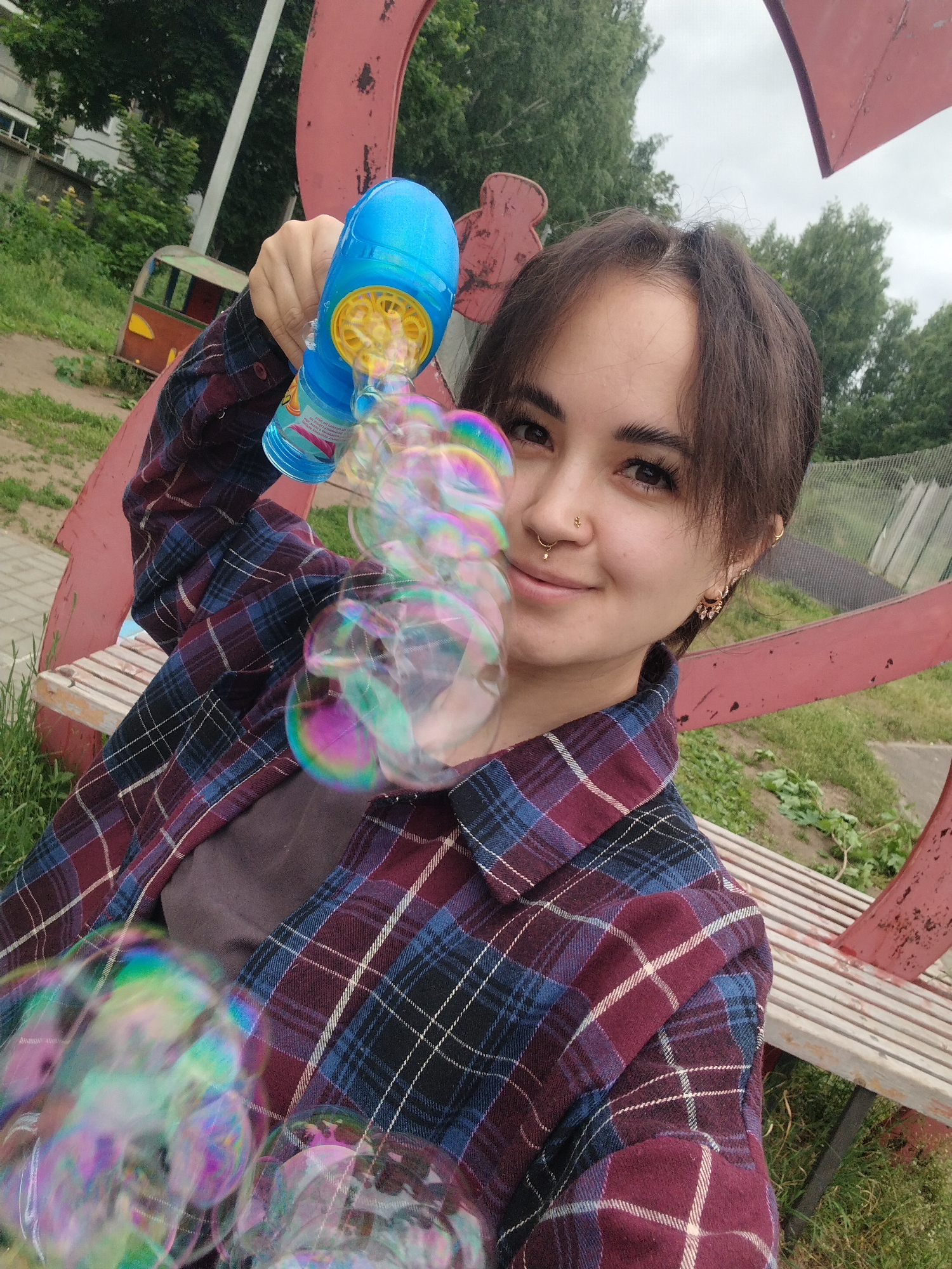 A young woman with dark hair is taking a selfie outdoors in Noviy, Russia. She is wearing a plaid shirt and holding a blue bubble gun that is producing a stream of iridescent bubbles. The woman is smiling and has a nose ring and earrings. In the background, there is a park-like setting with trees, a red metal structure, a wooden bench, and a small, colorful play structure resembling a miniature train car. The sky is overcast.