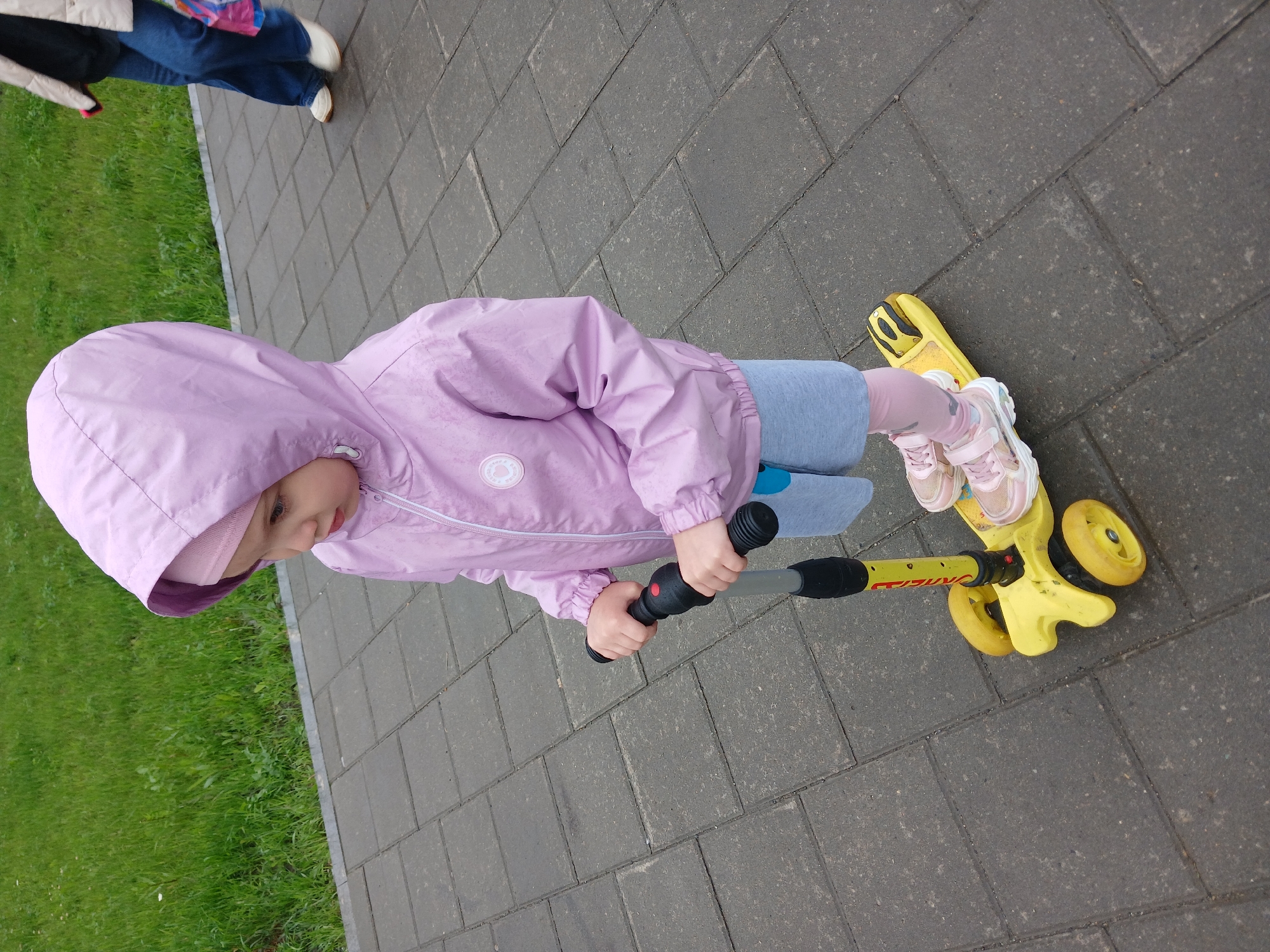 A young child stands on a yellow scooter on a paved sidewalk in Yekaterinburg, Russia. The child is wearing a light pink hooded jacket, a pink hat, light blue shorts, and pink and white sneakers. The child's hands are gripping the handlebars of the scooter. The scooter has a yellow platform and three yellow wheels. In the background, there is a grassy area and the legs of an adult are visible walking past. The pavement is made of large gray rectangular stones.