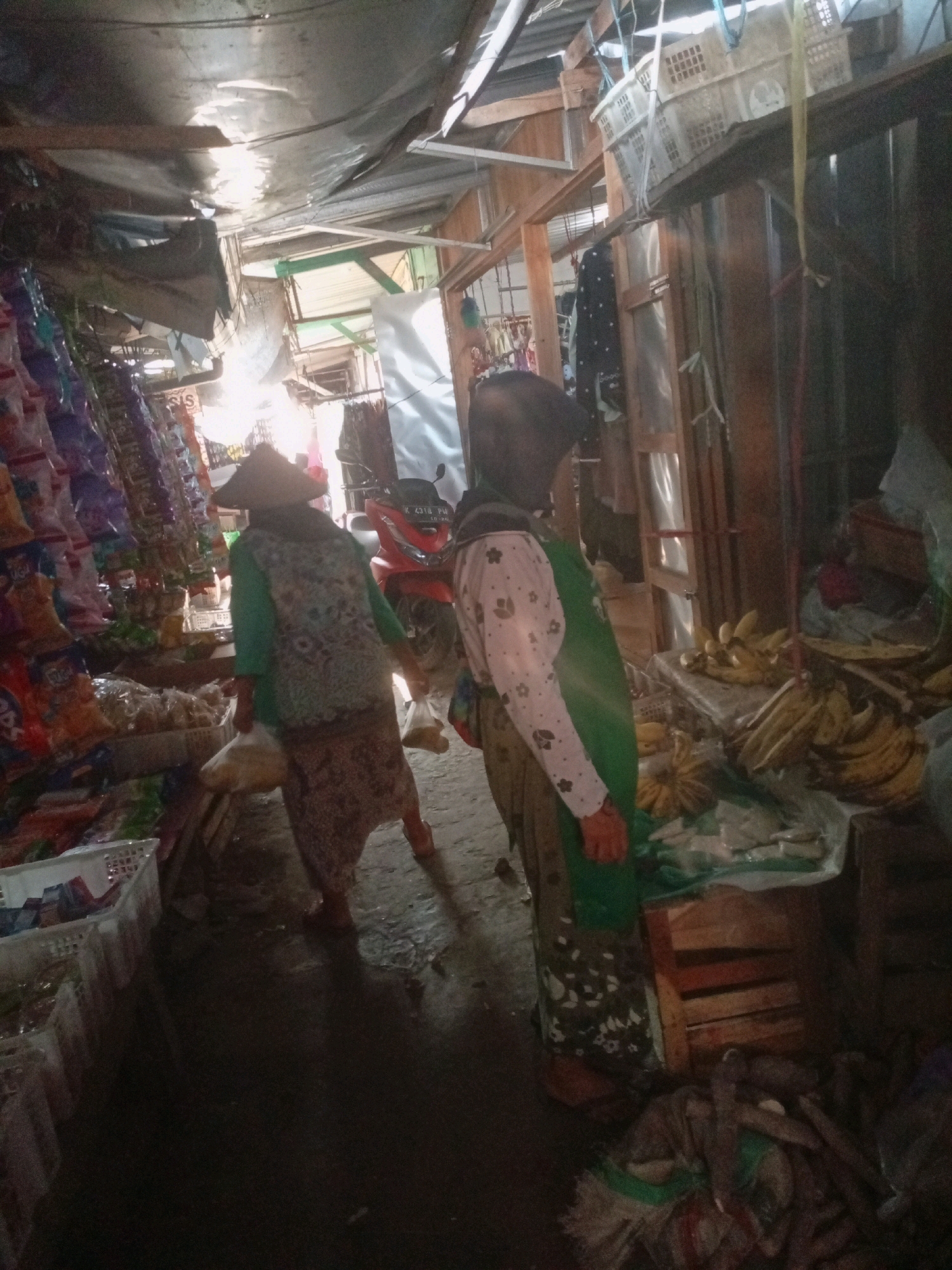 The image depicts a market scene in Sluke, Indonesia. Two women are visible, one wearing a conical hat and a green shirt over a patterned dress, carrying a plastic bag. The second woman is wearing a dark headscarf and a pink shirt with floral patterns, also with a green apron. She is standing near a stall laden with bananas. The market appears to be covered by a makeshift roof of corrugated metal and tarpaulin, creating a dimly lit environment. Stalls on the left display various packaged goods. The ground is wet, suggesting recent rain or cleaning. A red motorcycle is parked in the background. The overall impression is of a typical rural market in Indonesia.