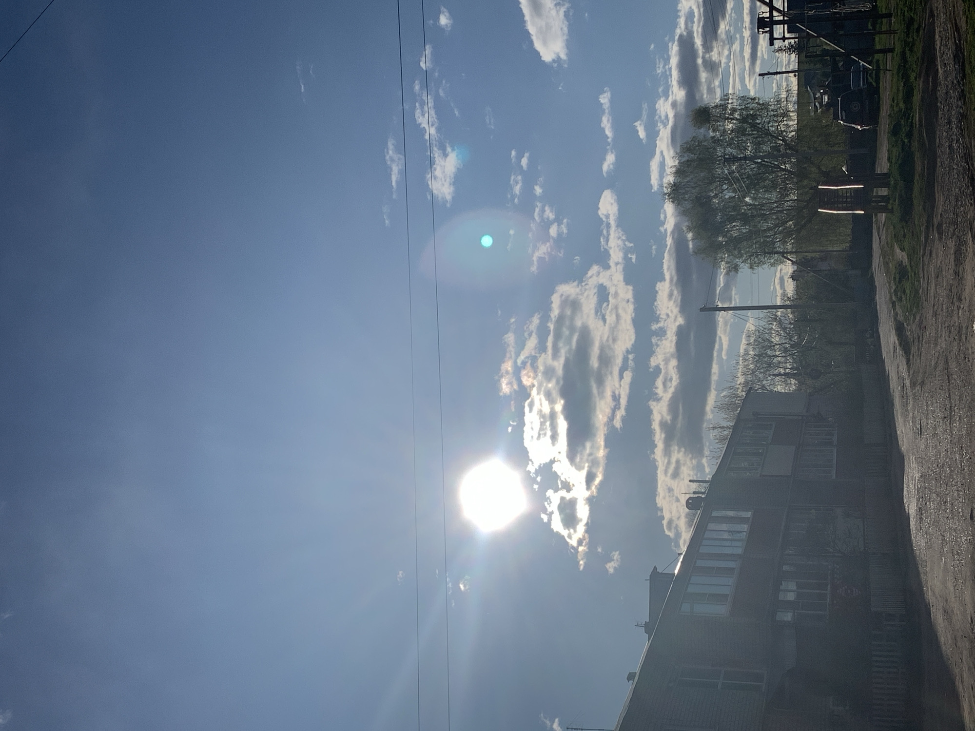 The image shows a bright, sunny day in what appears to be a residential area of the city of Фоки, Russia. The sun is high in the sky, partially obscured by scattered clouds, and its glare dominates the lower center of the frame. Power lines stretch across the blue sky. To the right of the frame, a multi-story residential building is visible, suggesting an urban or suburban setting. Further in the background, a treeline and utility poles can be discerned. The ground shows a narrow road or pathway along the right edge. No people are visible in the image.