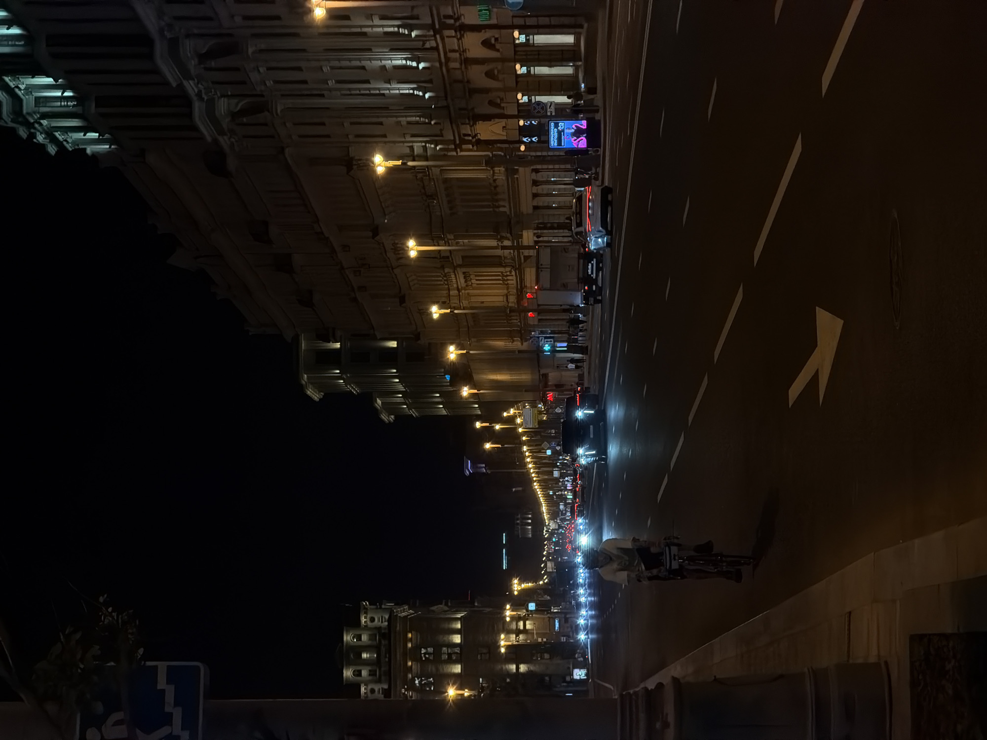 A street at night in Moscow, Russia. The road stretches into the distance with streetlights illuminating buildings on both sides. A person on a bicycle is on the right side of the road. There are cars in the distance. Road markings are visible, including an arrow pointing straight. The sky is dark.