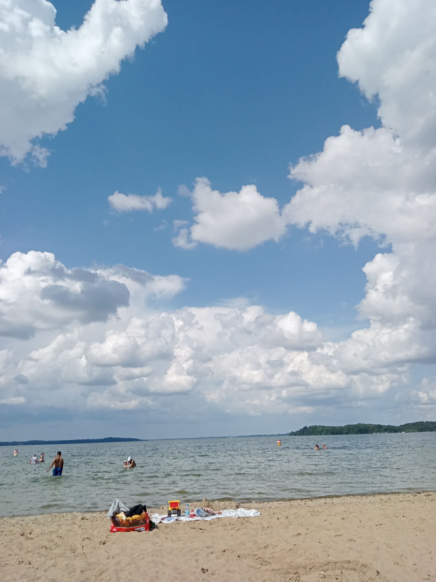 A bright, cloudy sky stretches above a wide, calm body of water. In the foreground, a sandy beach has several objects placed upon it. A red inflatable bag with items on top, including a yellow cooler, is positioned near the center. To the left, a white towel with a pattern is visible.
In the water, several individuals are visible. One person, wearing blue shorts, stands in the shallow water on the left side of the frame. Further out, multiple people are swimming or wading. A person in the middle of the frame is sitting on an object in the water, possibly a flotation device. In the distance, a line of trees marks the far shore of the lake, suggesting a natural landscape. A few buoys are also scattered across the water. The overall impression is a serene day at the beach.