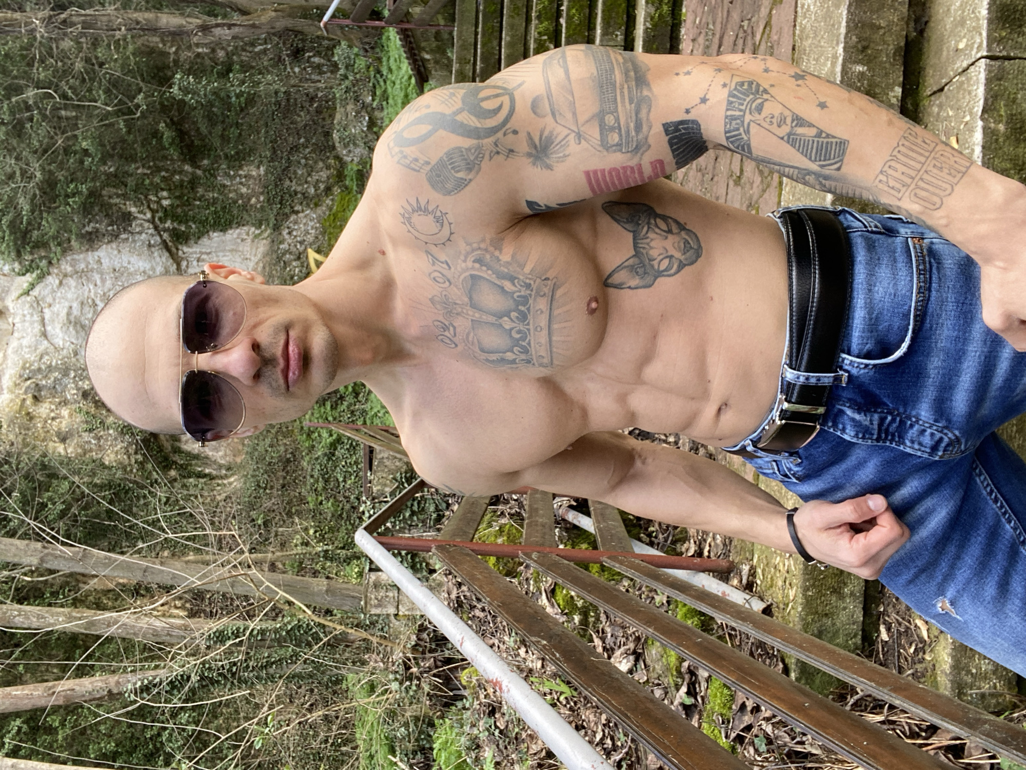 A bald, muscular adult male with dark aviator-style sunglasses stands shirtless, facing the viewer, in a vegetated, rocky outdoor area in Pleven, Bulgaria. He wears blue denim jeans with a distressed detail on the left thigh, a black leather belt with a silver buckle, and a black bracelet on his left wrist. Numerous black ink tattoos cover his chest and arms. On his chest, a large crown is centered, flanked by "02-07" and a crescent moon/sun motif. Below the crown, a sphynx cat head is visible. His right arm displays tattoos including microphones, musical notes, palm trees, a classic car, and the word "WORLD". His left forearm features a pharaoh's face integrated with the number "7", star constellations, and the text "GAME OVER". The subject's left arm is bent at the elbow with a loosely clenched fist. In the background, dense green foliage, bare tree branches, and a light-colored rock face are visible. A set of weathered stone steps ascends to the right behind the subject, accompanied by a wooden and metal railing with a silver-painted top rail and red supports on the left side of the frame. The ground is covered with dry leaves and moss.