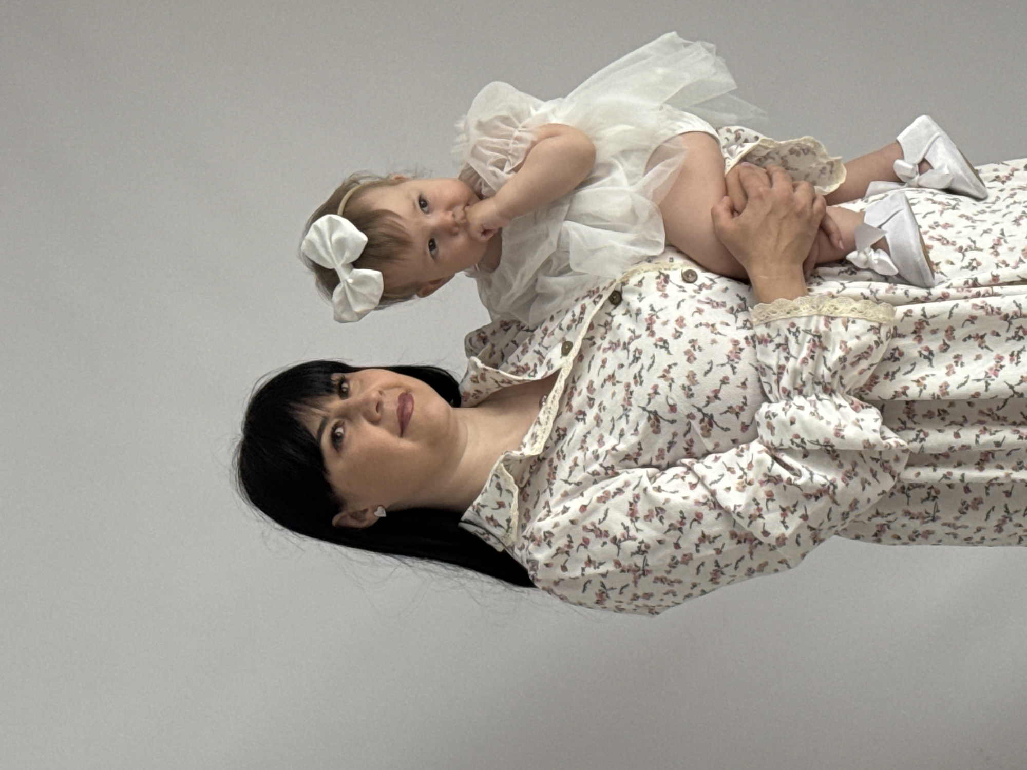 A medium shot shows a light-skinned woman lying on her back with a light-skinned baby lying on top of her. The woman has long, straight black hair and is wearing a white dress with small pink flowers and a lace collar. She looks up and to the left with a neutral expression. The baby is wearing a white dress, white bow headband, and white shoes with bows on them. The baby is also looking up and to the left with her finger in her mouth. The background is a solid light gray color. There are no visible indicators of the scene's location in the city of Izhevsk, Russia.