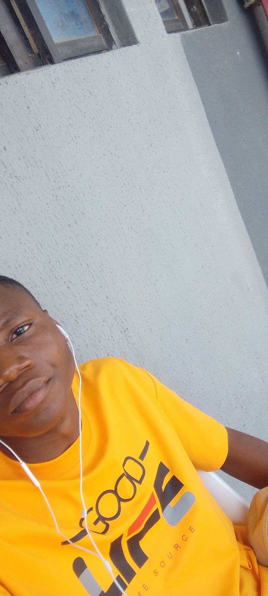 The image is a close-up, selfie-style shot of a young Black person wearing a bright yellow t-shirt with a graphic design and white earphones. The person is looking towards the camera with a slight smile, their head tilted to the side. The t-shirt has bold, stylized lettering and shapes in black, grey, and red. Part of the text "LIFE SOURCE" is visible at the bottom of the graphic.
The background consists of a textured, light grey wall, possibly stucco, with a shadow cast diagonally across it. Above the wall, in the top left corner, a sliver of a window or opening can be seen, with dark horizontal bars and a bluish tint, suggesting it might be a window with metal bars or a ventilation grate. The lighting suggests it is daytime, and the overall scene appears to be outdoors or in a well-lit, open area. The setting seems casual and relaxed. The individual's expression is pleasant and engaging.