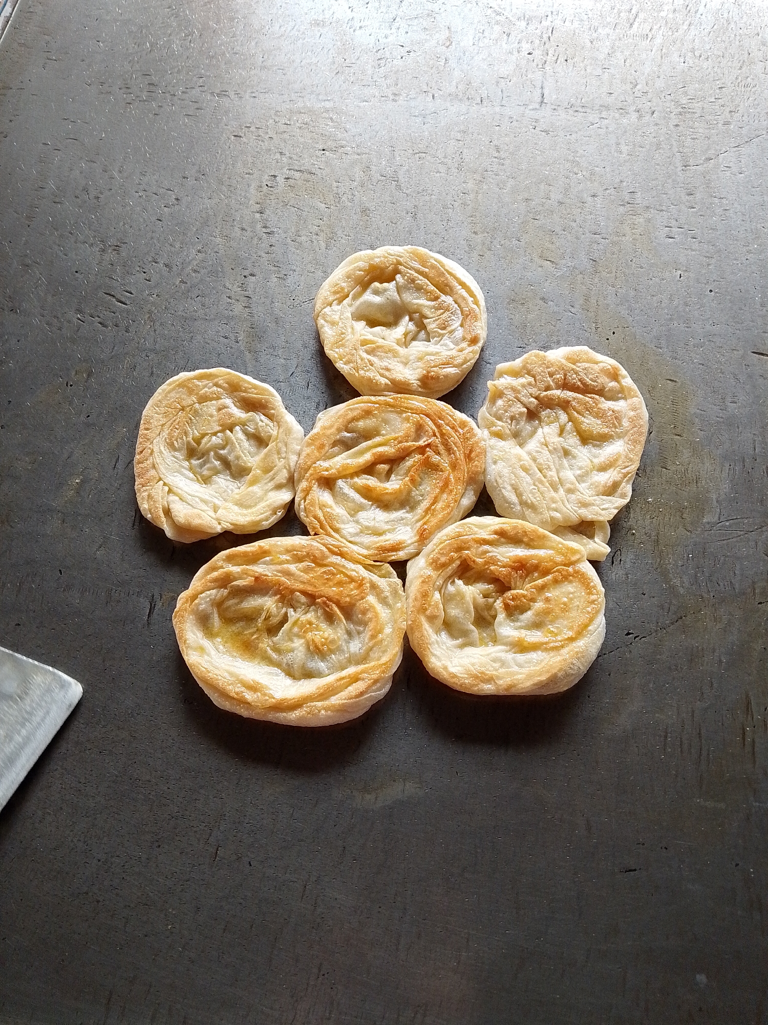 Six circular, flaky pastries with golden-brown tops are arranged on a dark, textured surface that appears to be a metal tabletop. The pastries have a layered, coiled appearance, suggesting they are a type of flatbread or pastry. Some of the pastries have a slightly oily sheen. In the bottom left corner of the image, a portion of a metallic object with a flat edge is visible, possibly a spatula or a knife. The background surface has a worn, industrial look with visible scratches and a mottled texture. There are no people or animals present in the image. The lighting suggests an overhead light source casting subtle shadows.
The image does not contain any information that would allow for a specific location within Lhokseumawe, Indonesia, or any other city.
