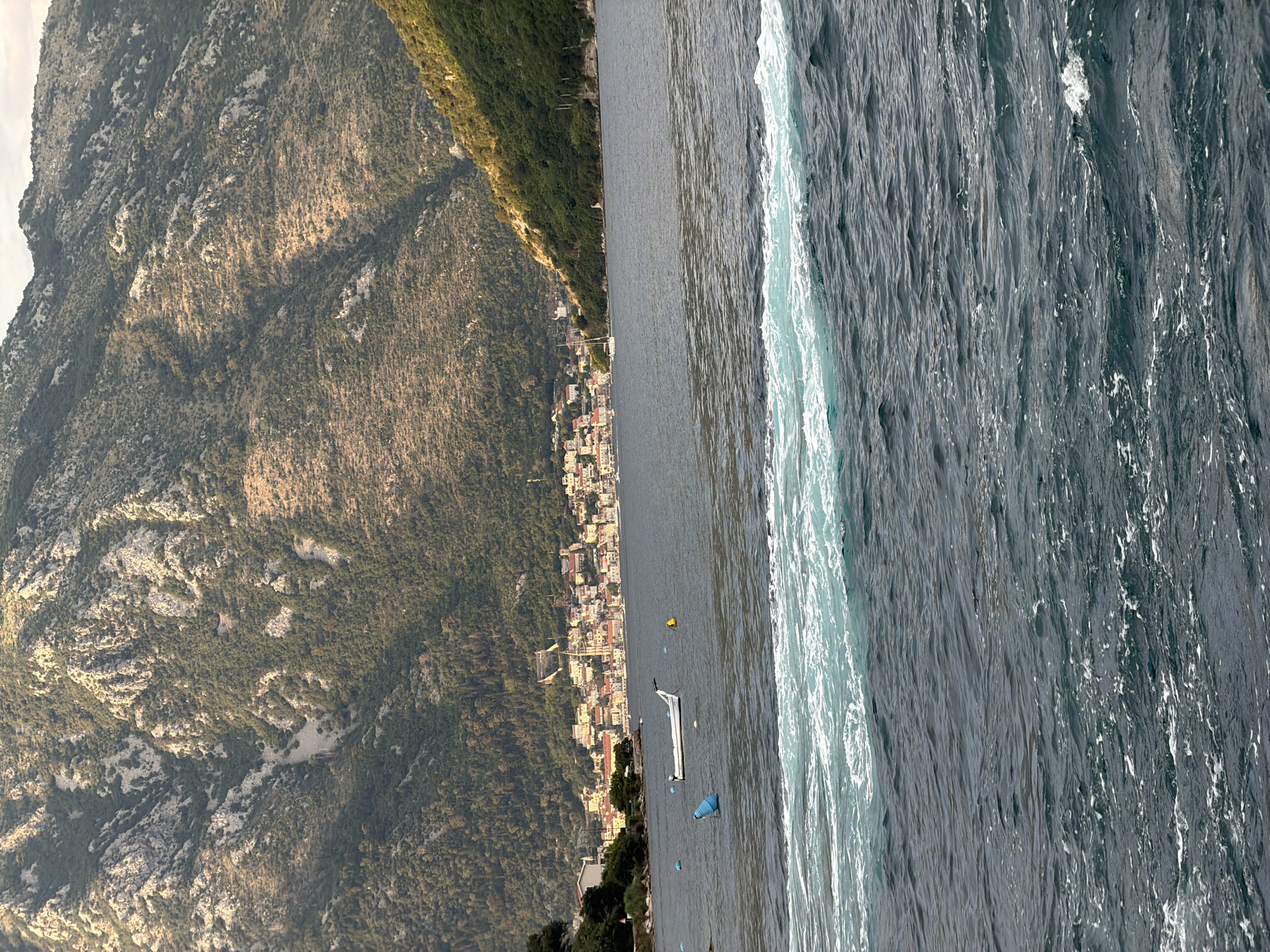 This is a medium shot photograph taken from a body of water looking towards a coastal town situated at the base of a steep, mountainous incline. The water in the foreground is choppy with significant white foam and a distinct band of turquoise-colored turbulent water running horizontally across the frame.
In the middle ground, the dark blue water is calmer, with a white motorboat visible in the center. Two buoys, one blue and one yellow, are also present in the water. To the left of the boat, a white structure, possibly a building, is partially visible on the shore.
The background features a densely populated coastal town with buildings exhibiting red-tiled roofs and light-colored facades. The town is nestled against a large, rugged mountain that dominates the upper portion of the image. The mountain's slopes are covered with sparse vegetation and rocky outcrops. The lighting suggests it is either early morning or late afternoon, as the sun casts shadows across the mountain.
The scene appears to be in or near Đurići, Montenegro, given the characteristic landscape of the Bay of Kotor, which features steep mountains meeting the sea and picturesque coastal towns. There are no people clearly visible in the image.