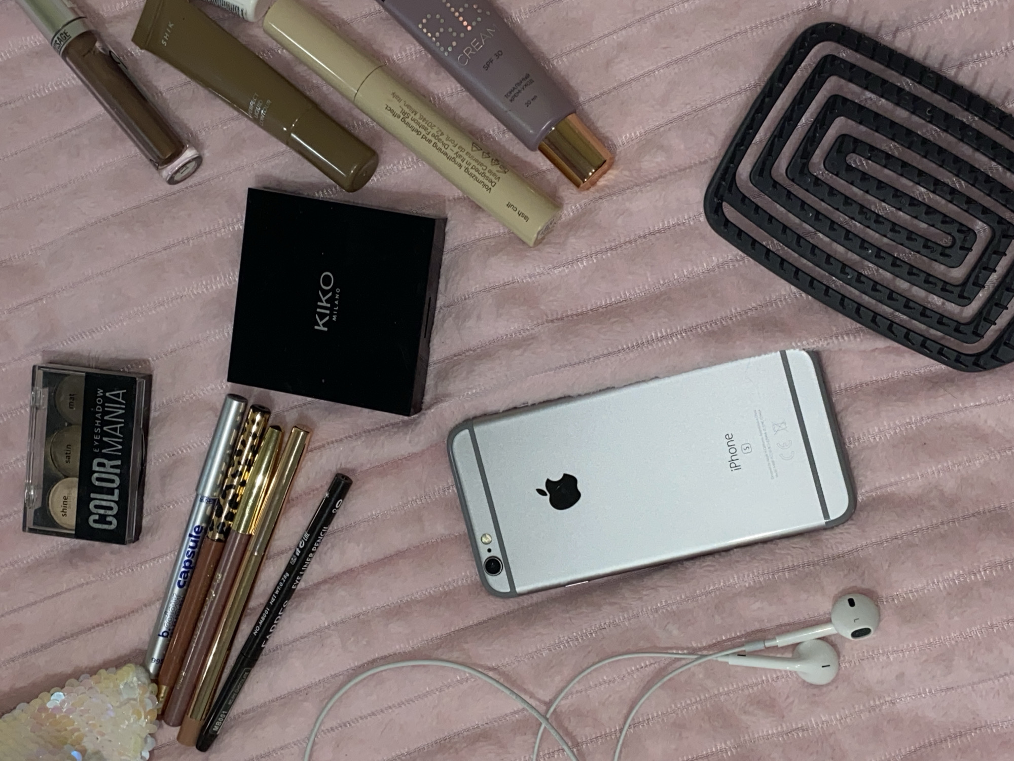 The image displays a flat lay arrangement of various cosmetic products and personal electronics on a textured light pink surface, likely a blanket or towel. No people are present.
In the upper left section, there's a dark brown liquid cosmetic container with "SAGE" visible. Adjacent to it, a dark olive-brown tube is labeled "SHIK PERFECT LIQUID CONTOUR." Further right, a light beige tube reads "lash cult" and provides manufacturing details: "Volumizing, lengthening and defining effect. Designed in Italy - Divage Fashion SRL. Viale Caterina da Forli, 42, 20146, Milan, Italy." Next to this, a purple tube states "CREAM SPF 30" and "ТОНАЛЬНЫЙ КРЕМ-УХОД" (Tonal Cream-Care) with "20" (likely ml).
A black square compact branded "KIKO MILANO" is centrally located on the left. Below it, a clear plastic eyeshadow palette features four shades (shine, satin, mat) and is labeled "EYESHADOW COLOR MANIA capsule." A group of pencil-style cosmetics, including shades of silver, bronze, and gold, are next to the eyeshadow palette. A black pencil is identified as "CADDES EYE LINER PENCIL" with "NO M8001 NET WT:0.28g."
A silver iPhone, identifiable as an 'S' model (likely 6S), rests on the right side, showcasing its Apple logo and "iPhone S" along with CE markings. White wired Apple earphones are positioned below the phone.
In the upper right corner, a black silicone mat with a distinctive rectangular spiral design is visible. A small cluster of iridescent sequins is in the bottom left corner.
The background is a soft, ribbed pink fabric. The presence of Cyrillic text ("ТОНАЛЬНЫЙ КРЕМ-УХОД") on one cosmetic item is consistent with the provided location context of Votkinsk, Russia. The image is a static shot, offering no indication of time of day or activity.