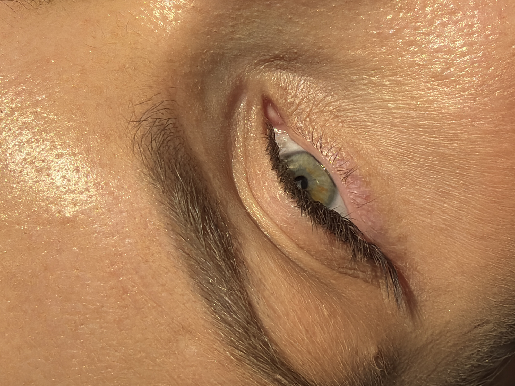This is a close-up, high-angle shot of a person's left eye and surrounding forehead area. The skin appears to have a dewy or glittery sheen. The eye is open, revealing a light green iris with dark brown eyelashes and eyebrows. The eyelid is subtly lined with dark makeup. The lighting casts a shadow across the upper part of the forehead, highlighting the texture of the skin. There are no identifiable landmarks or context that would place this image in Moscow, Russia, or any other specific geographic location.