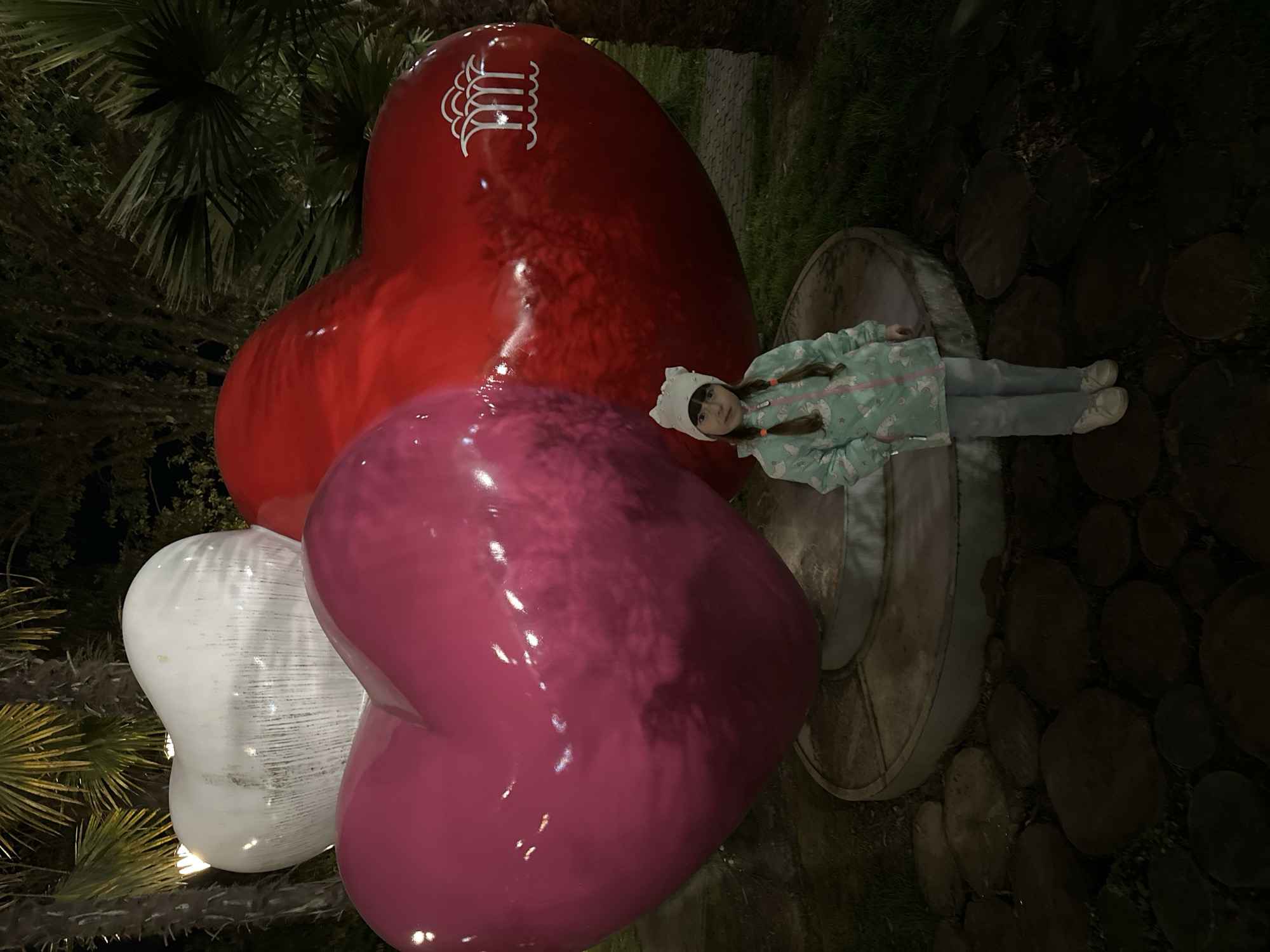 A nighttime outdoor scene captures a young child lying on her back next to a playful display of three oversized heart sculptures. The child, who appears to be a girl, is dressed in a light blue jacket adorned with a unicorn pattern, light-colored pants, and a white knitted hat with small ear-like details. She has her hair styled in two pigtail braids and looks towards the camera with a calm, neutral expression.
The prominent heart sculptures are rendered in vibrant colors and a glossy finish, reflecting light. The largest heart is red and positioned at the top, featuring a distinct white stylized "M" logo on its upper surface. Below it is a bright pink heart, and to its left, a white, translucent heart is visible, showing internal illumination.
The surrounding environment is dark, suggesting evening or night, with glimpses of lush, dark green palm-like foliage in the background and foreground, indicative of a subtropical setting. To the child's right, a circular, low structure, possibly a planter or bench, is visible. The ground is covered with dark, round wooden logs, forming a rustic path or decorative surface. The overall impression is a whimsical encounter with public art in an illuminated garden or park setting, consistent with the locale of Sochi, Russia.