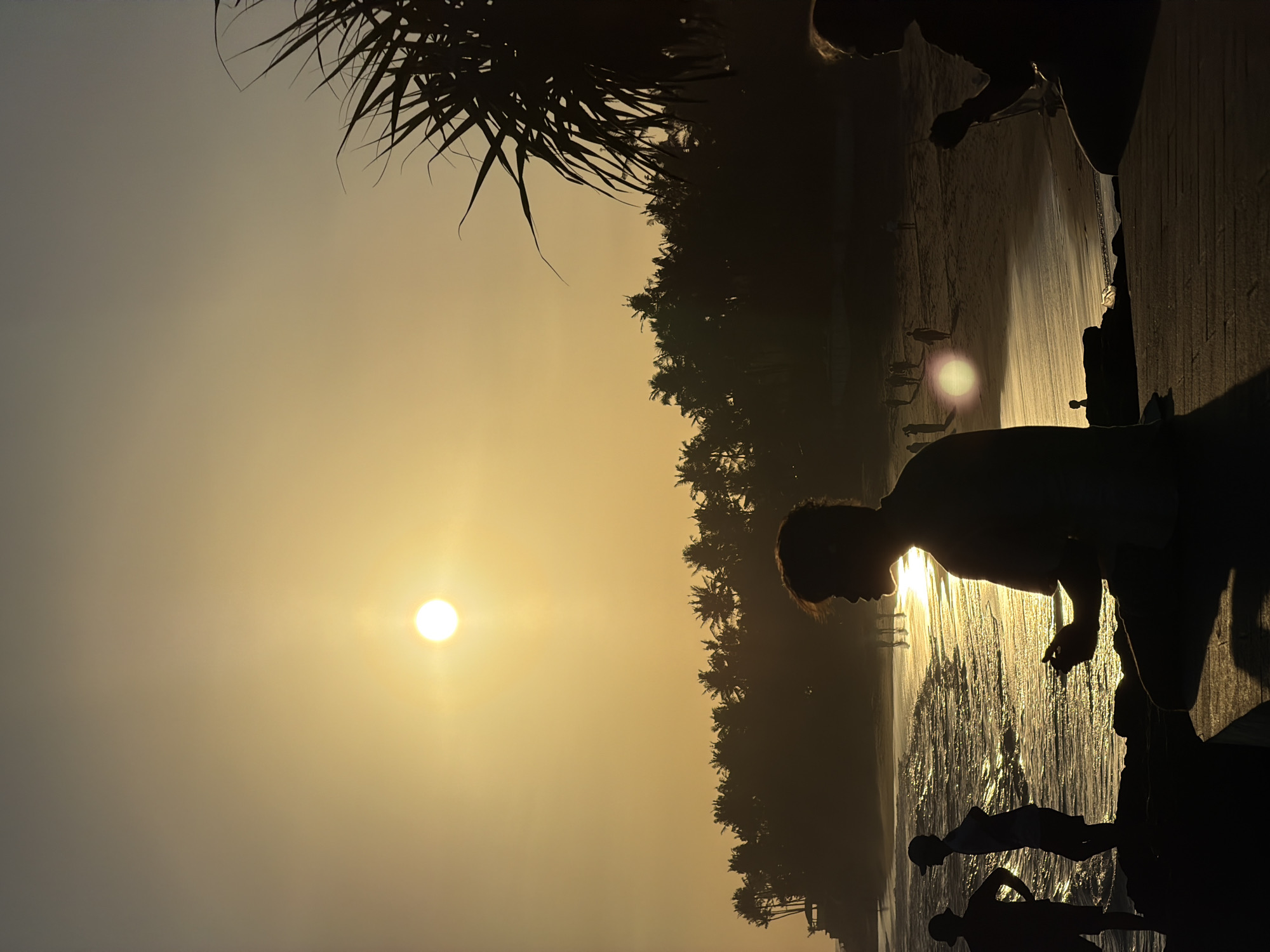 This media file captures a serene tropical beach scene at either sunrise or sunset, likely in Mengwi, Indonesia. The sun, a bright, circular golden orb, is low in the sky towards the left, bathing the entire landscape in a warm, radiant glow. The sky exhibits a gradient of soft orange and yellow hues, with the upper portion appearing hazier.
Several palm trees are silhouetted against the brilliant light, with their fronds visible prominently at the top edge and forming a dense, dark line along the distant horizon, bordering the sandy beach. The ocean's surface reflects the sun's brilliance, creating a shimmering, golden path on the water that reaches towards the foreground.
Numerous human figures are visible, depicted as dark silhouettes against the bright backdrop. In the foreground, a prominent male figure stands near the water's edge, seemingly looking down or out towards the sea. Further along the wet sand, additional smaller figures, possibly including children, are also present, their forms indistinguishable in the strong backlighting. The activity appears to be people enjoying the tranquil beach environment during twilight. The image conveys a peaceful and picturesque atmosphere, characterized by strong backlighting and dramatic silhouettes. No visible text is present.