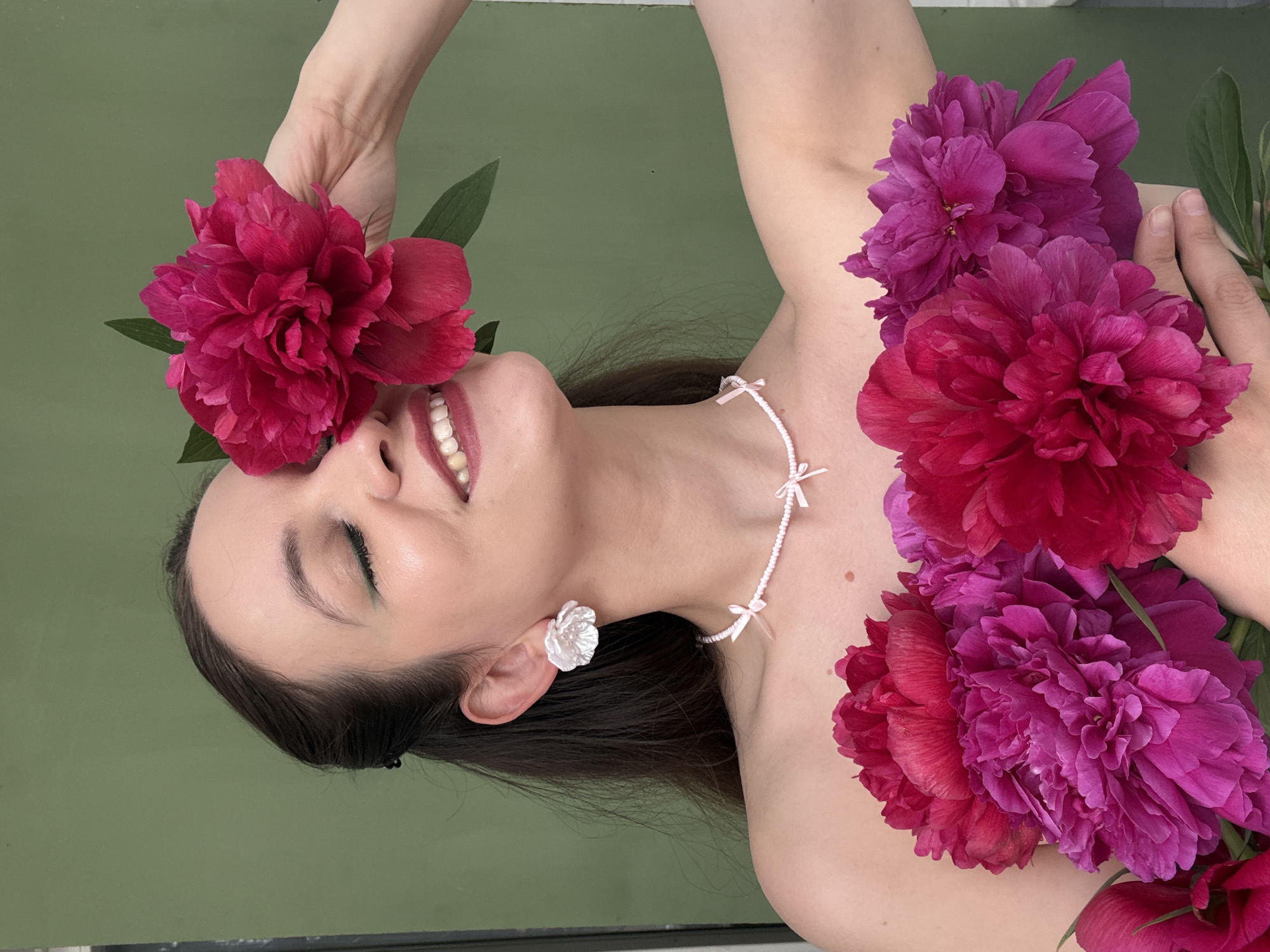 The image shows a woman against a muted green background. She is holding a bright pink peony to her face, with her eyes closed and a smile on her lips. She is wearing a delicate white beaded necklace with small pink bows, and white flower-shaped earrings. Several other peonies in shades of pink and magenta are arranged in front of her chest. Her hair is dark and pulled back, with a few strands loose around her face. The lighting is soft, highlighting her skin and the vibrant colors of the flowers. There is no indication of the city of Chaikovsky, Russia in the image.