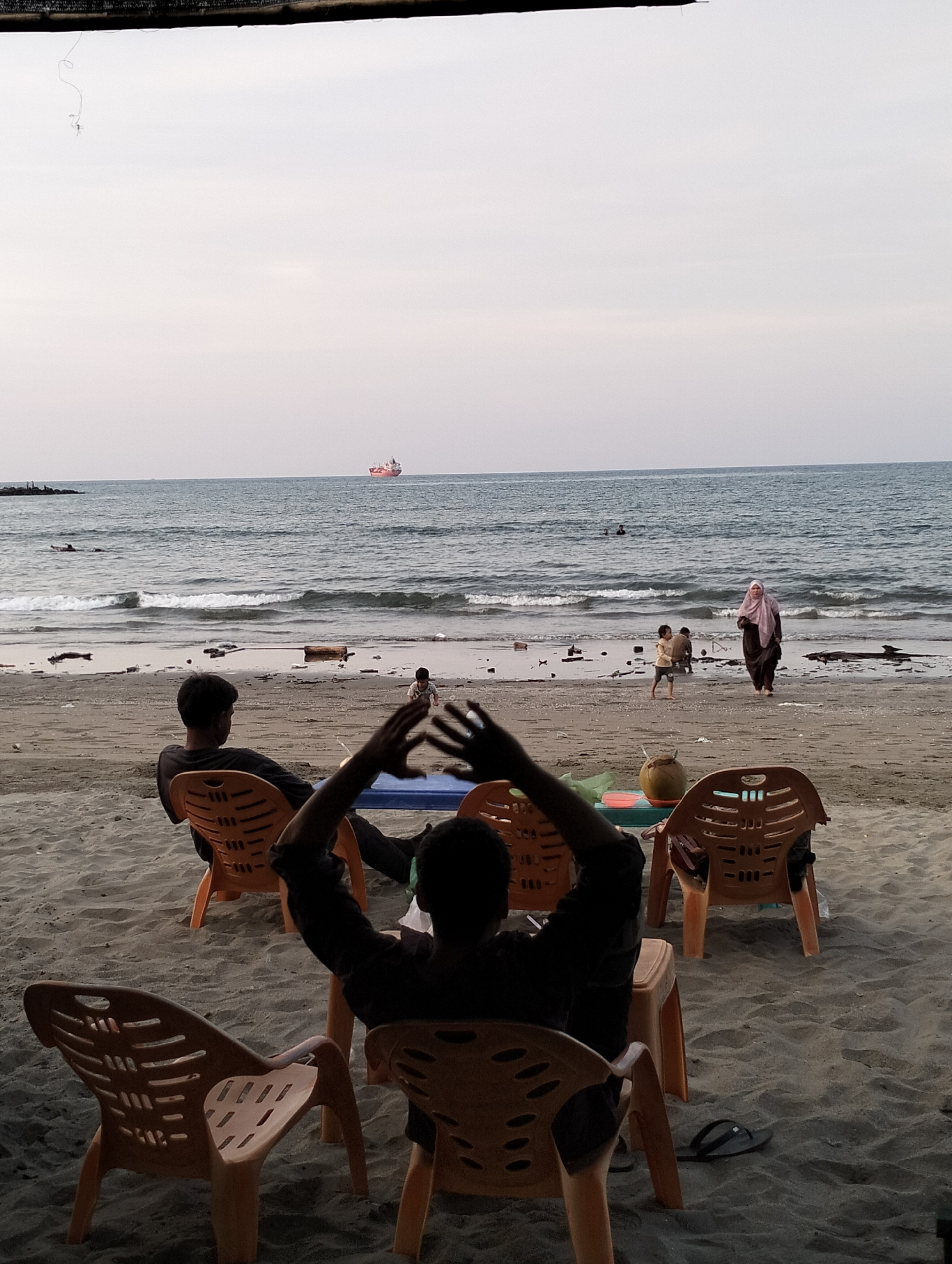 This image captures a tranquil beach scene in Lhokseumawe, Indonesia, likely during late afternoon or early evening under an overcast or hazy sky. The overall atmosphere is calm and relaxed.
In the foreground, several individuals are seated on orange plastic chairs directly on the sandy beach, facing the ocean. One person in the immediate foreground is silhouetted, with their hands raised, possibly stretching or gesturing. To their left, another person is also silhouetted, seated and looking out at the sea. An empty orange plastic chair is visible in the bottom-left. A small blue table, holding a green coconut and other small items, is positioned in front of some of the chairs.
The midground shows the sandy shoreline where gentle waves break. A woman wearing a pink hijab and a long dark dress walks along the waterline, accompanied by a young child. Another child and an adult are further along the beach, engaged in casual activities. Scattered debris or natural elements are visible on the wet sand.
In the background, the vast ocean extends under the soft, diffused light. A red and white cargo ship or tanker is visible on the distant horizon, indicating maritime activity. The water is calm with small ripples and breaking waves near the shore. At the very top of the frame, a dark, blurry structure, possibly an awning or roof, is visible, from which a thin wire or string dangles.
No discernible text is visible in the image. The scene suggests a moment of leisure and observation by the coast.