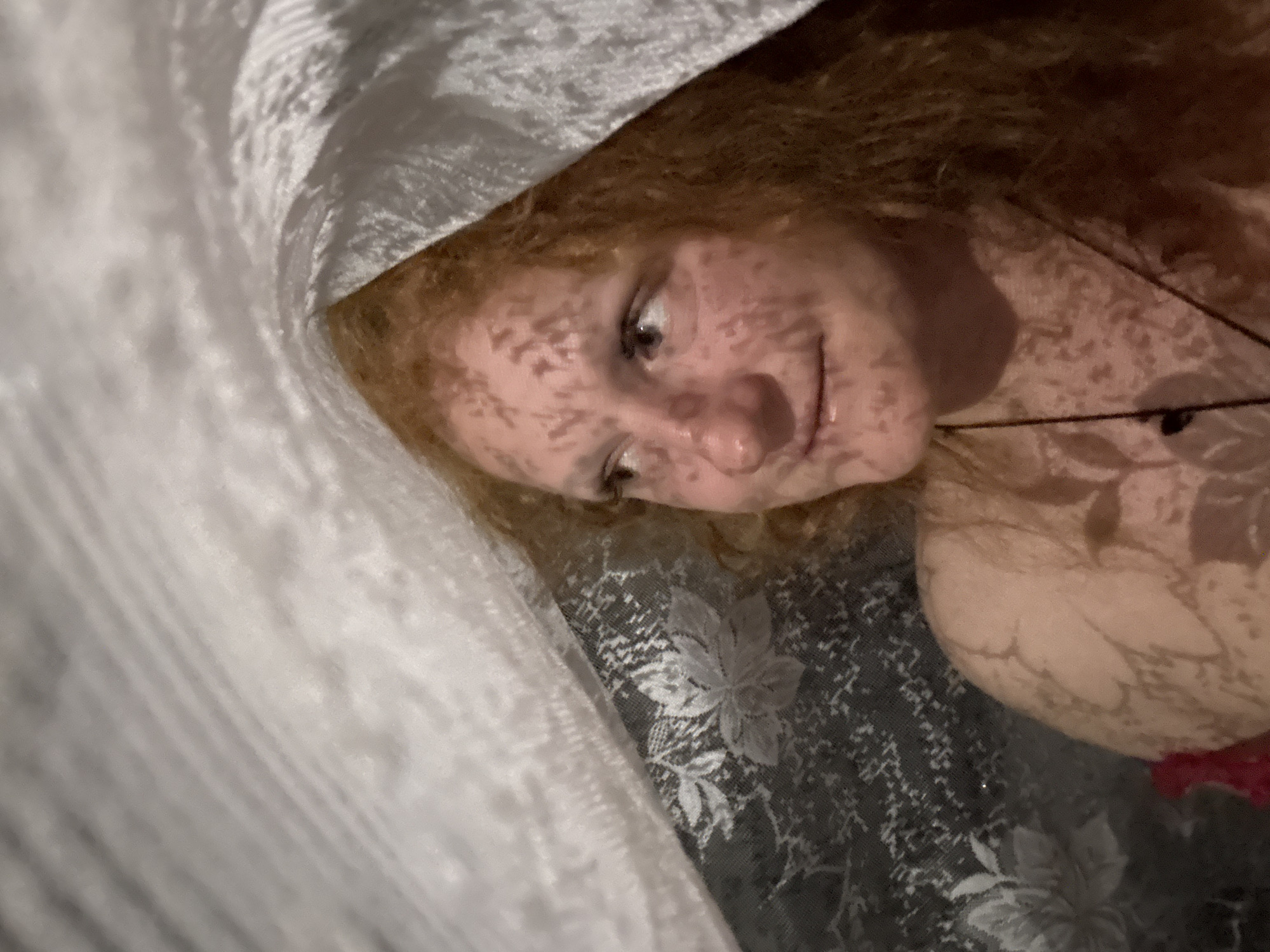 A close-up indoor shot depicts a person with fair skin and red hair. The person's face is partially covered by a patterned shadow, likely from lace or a similar material. The person is smiling slightly and looking to the left. Part of a white, possibly sheer fabric is draped over the person's head. The background features a floral patterned fabric. There is no indication of this image being taken in the city of Chaikovsky, Russia.