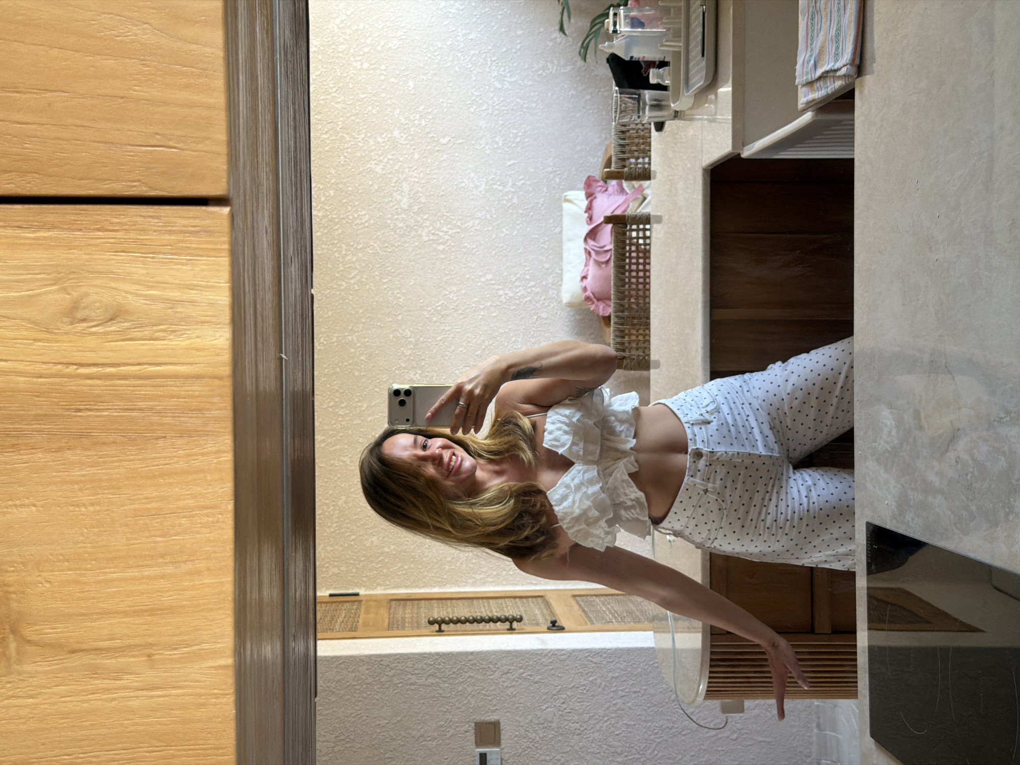 A woman is taking a selfie in a bathroom mirror. She is wearing a white ruffled crop top and white pants with dark polka dots. Her hair is brown and shoulder-length. The background of the mirror shows a bathroom with light-colored textured walls, wooden cabinets, and a sink. A woven basket with pink pillows is visible on a counter. The location is identified as Mengwi, Indonesia.