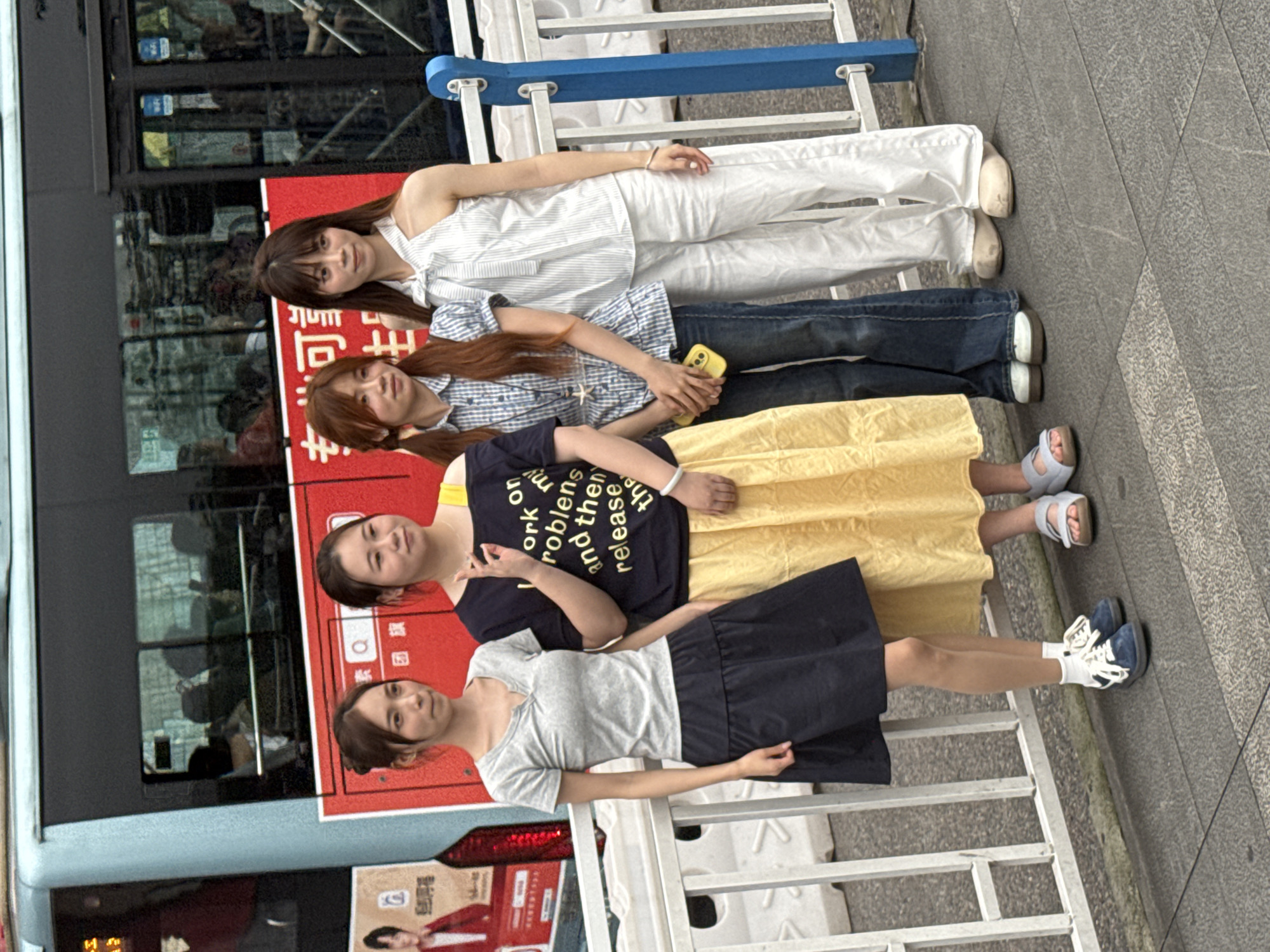 Four Asian women are standing in a row in an urban environment, likely Yuzhong District, China, based on visual cues. The women are of varying heights and are dressed in casual clothing. The first woman, positioned at the top, is wearing a white top and white pants. The second woman has on a blue and white checkered shirt and jeans. The third woman is wearing a dark-colored t-shirt with white text and a yellow skirt. The fourth woman at the bottom is wearing a gray t-shirt and a navy blue skirt, with white socks and blue and white sneakers. Behind the women is a parked blue bus. A red sign is visible on the side of the bus. The women are standing on a gray sidewalk next to a metal railing. The background includes hints of a busy urban street.