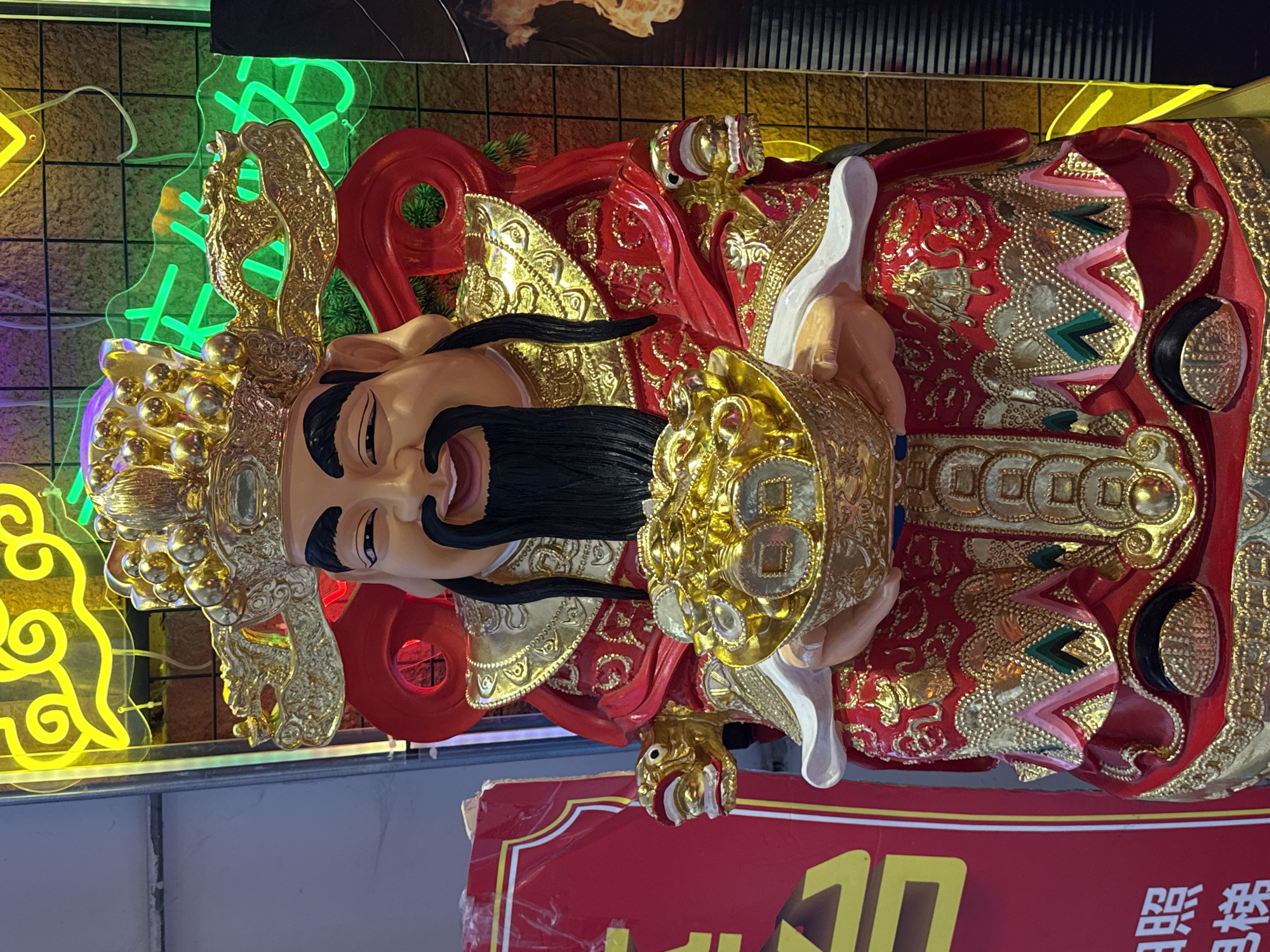 A medium shot, taken indoors in Yuzhong District, China, depicts a red and gold statue of a deity with a long black beard and mustache, smiling, eyes closed. The statue is richly decorated and wears a golden crown. The deity holds a golden bowl full of coins. Behind the statue are neon lights in various shapes and colors. The bottom part of a red sign with text is also visible.