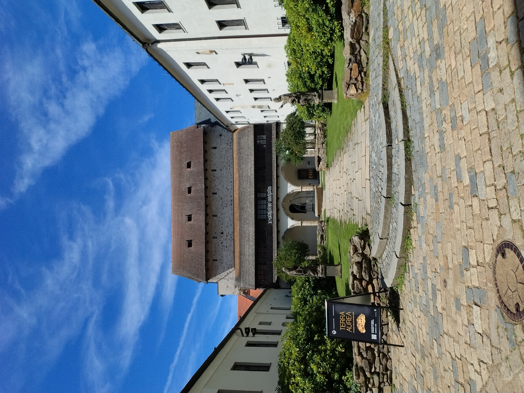 The image depicts a courtyard of a historical building, likely part of the Písek Castle complex in Písek, Czechia. The courtyard is paved with cobblestones. In the foreground, to the left, is a black A-frame sign with text and an image; it reads "TERRA AUREA" and appears to be an advertisement or informational display. The courtyard leads to a walkway with stone steps and then a gravel path. On either side of the path are manicured green bushes and two stone statues. The main building in the background features a stone tower with a tiled roof and an arched walkway on the ground floor. White buildings with many windows flank the courtyard on the left and right. The sky above is bright blue with scattered white clouds. The overall scene suggests a sunny day in a historic location.