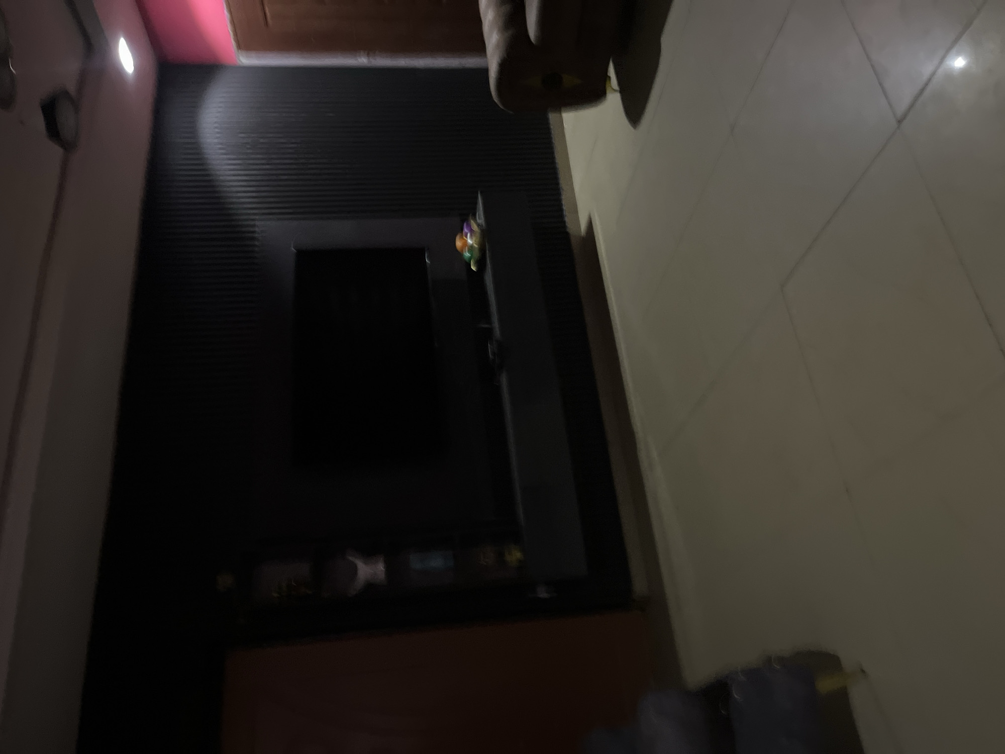 The image shows a dimly lit living room, likely in Jos, Nigeria. A large black television is mounted on a fluted black wall. Below the television is a dark shelf displaying several colorful decorative balls. To the right of the television, part of a brown leather sofa is visible. To the left of the television, a partially visible dark purple chair or ottoman with yellow legs is present. A pink painted wall and a wooden door are visible on the far right of the frame. The floor is tiled in a light color, and there is a ceiling fan visible at the top of the image. The overall impression is of a modern, sparsely decorated living space.