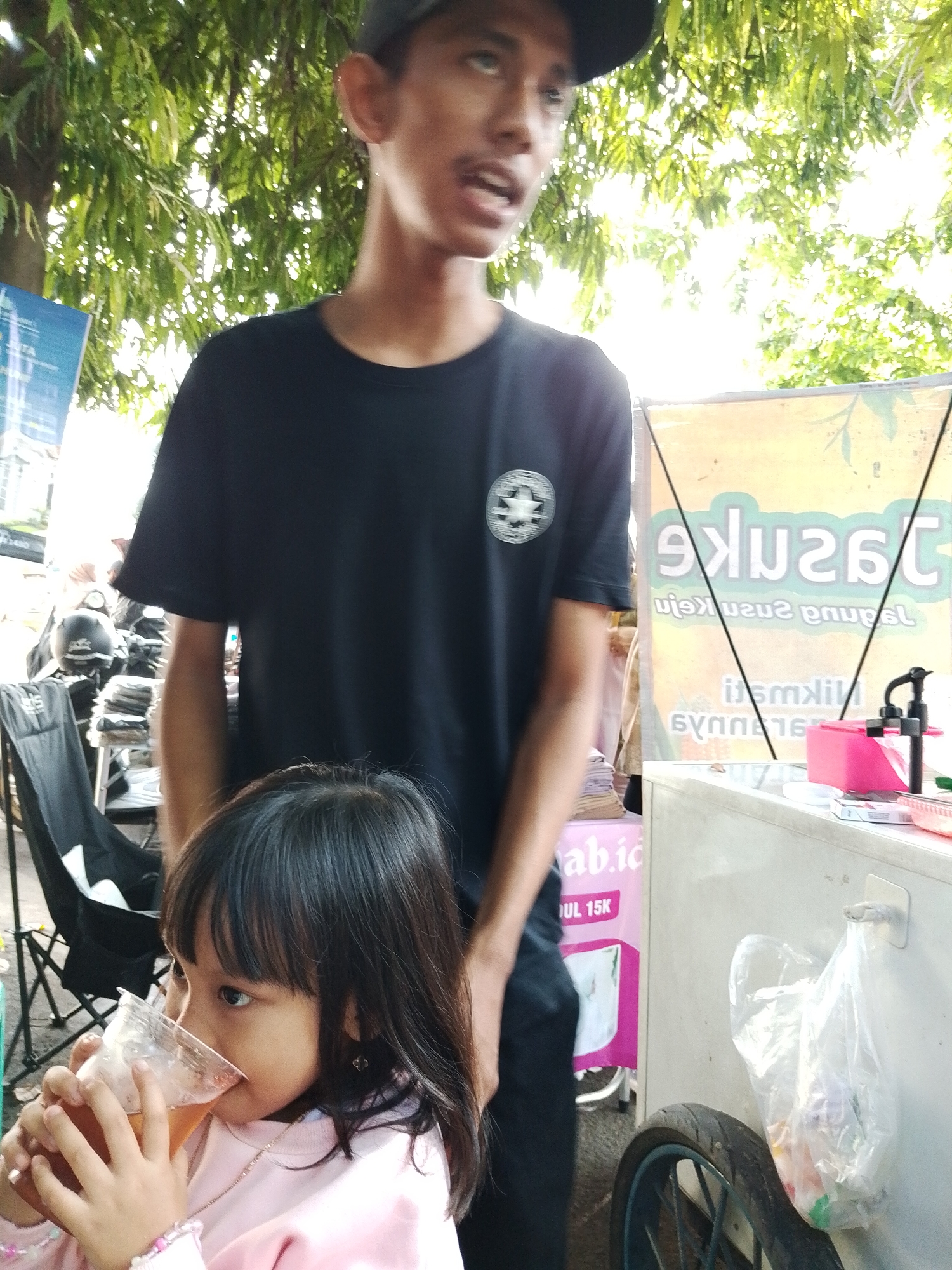 The photograph captures a scene in Kecamatan Pati, Indonesia, featuring a young girl drinking from a clear plastic cup in front of an adult male. The girl is wearing a light pink sweater. The man is standing behind her, dressed in a black t-shirt with a logo and a black baseball cap. He is looking off to the side and his mouth is open as if he is speaking. Behind the man, there is a sign with Indonesian script. A beverage cart or stand is to the right, with a plastic bag hanging from its side. To the left, there's a black folding chair. The setting is outdoors, with trees visible overhead.