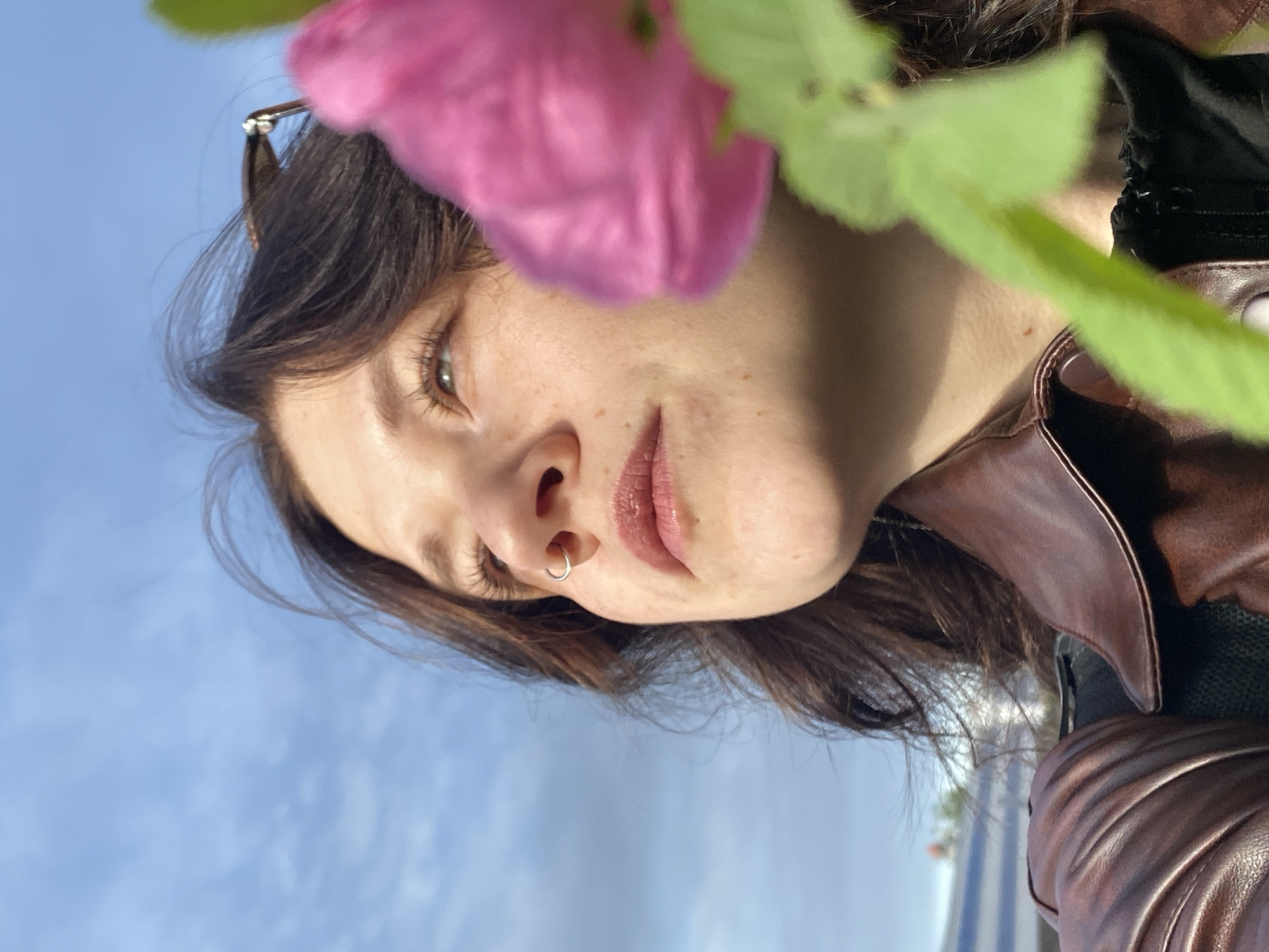 A low-angle shot depicts a woman with brown hair, a nose ring, and a brown leather jacket. A pink flower and green leaves are partially obscuring the view. The background shows a blue sky with white clouds. A building and a small strip of land are visible in the distance, suggestive of the city of Chaikovsky, Russia.