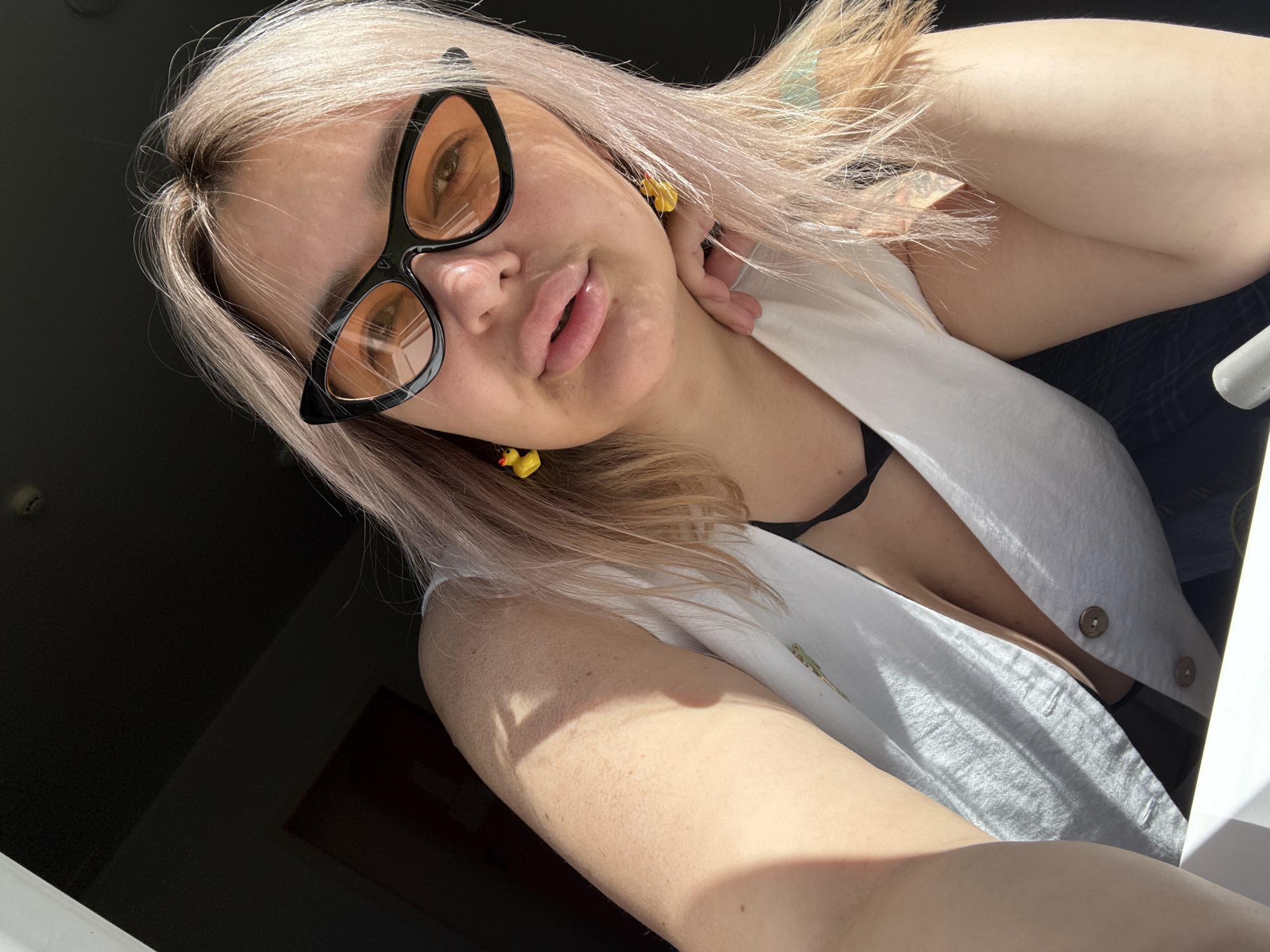 A close-up, angled selfie shows a woman with light blonde and pink-tinted hair, wearing cat-eye sunglasses with orange lenses and duck-shaped earrings. She is wearing a white V-neck vest over a black bra, with the buttons undone, exposing her décolletage. The lighting suggests she is near a window, with sunlight casting shadows on her arm and shoulder. The background is dark, with a hint of a doorway visible on the left. The scene is indoors, and no specific location in Birobidzhan, Russia, can be identified.