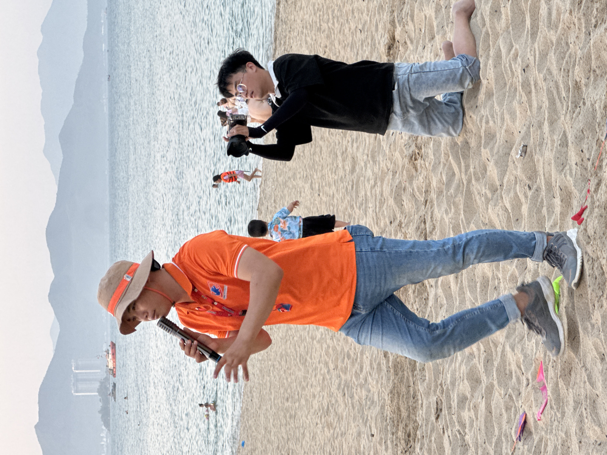 The image depicts a sandy beach with the ocean and mountains in the background. In the foreground, a man wearing an orange polo shirt, blue jeans, and a bucket hat is holding a microphone and speaking. He appears to be addressing someone or a group of people. To his right, a young man wearing a black long-sleeved shirt, glasses, and grey shorts is kneeling and taking a photo with a camera. Several other people are visible in the background, some in the water and others on the beach. There are boats and buildings visible on the horizon. The scene suggests a public event or gathering taking place on a beach, possibly in Nha Trang, Vietnam, given the presence of the ocean, mountains, and what appear to be urban structures in the distance. The atmosphere appears to be casual and possibly recreational.