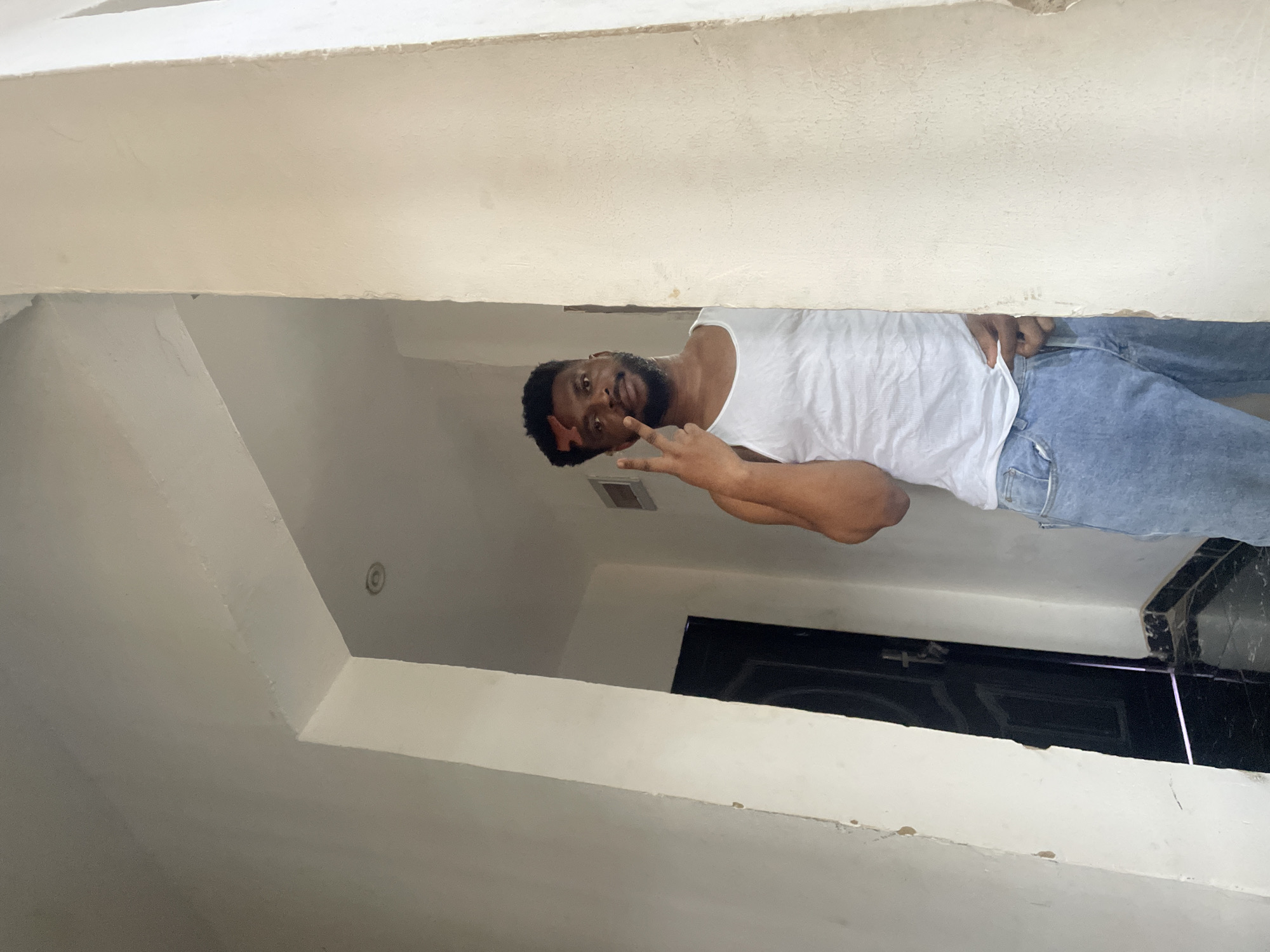 A man with dark hair and a beard, wearing a white tank top and blue jeans, is positioned in a doorway or passage. He is giving a peace sign with his right hand. The environment appears to be indoors, with white walls and ceilings. In the background, there is a black door. The location is possibly in Abuja, Nigeria.