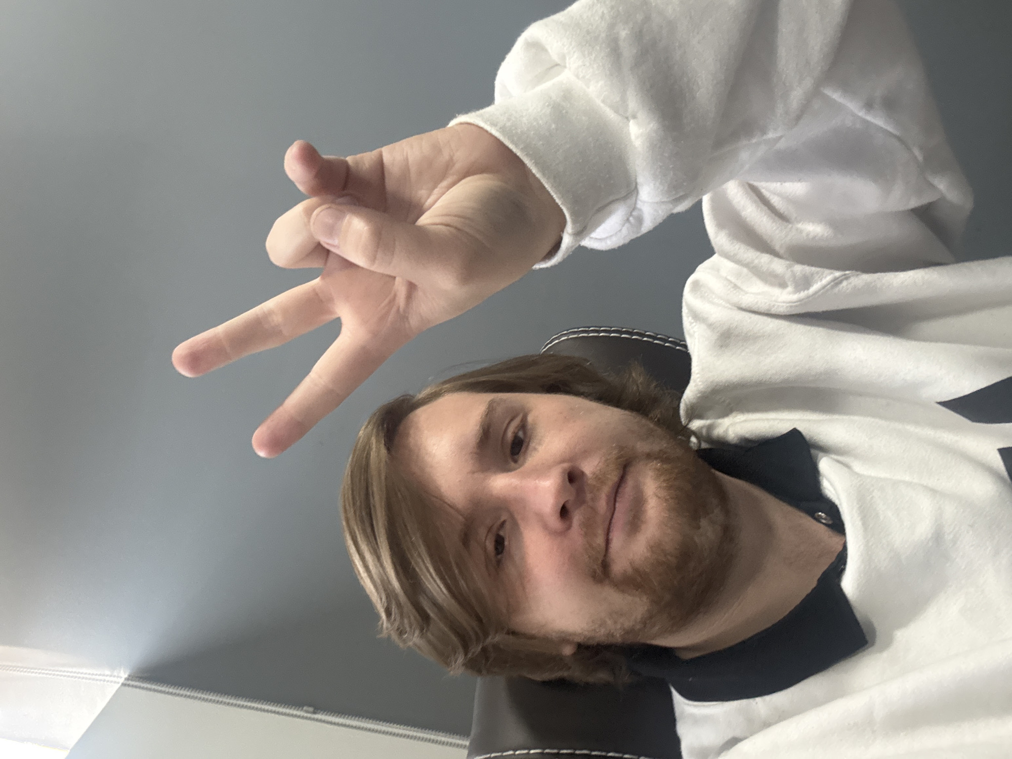 A selfie-style image shows a fair-skinned individual with shoulder-length blonde hair and a reddish beard. The person is wearing a white sweater with a black collar peeking out from underneath. A hand, also fair-skinned and wearing the same white sweater, is making a three-finger gesture above the person's head. The background is a plain grey wall. The location is not possible to determine from this image.